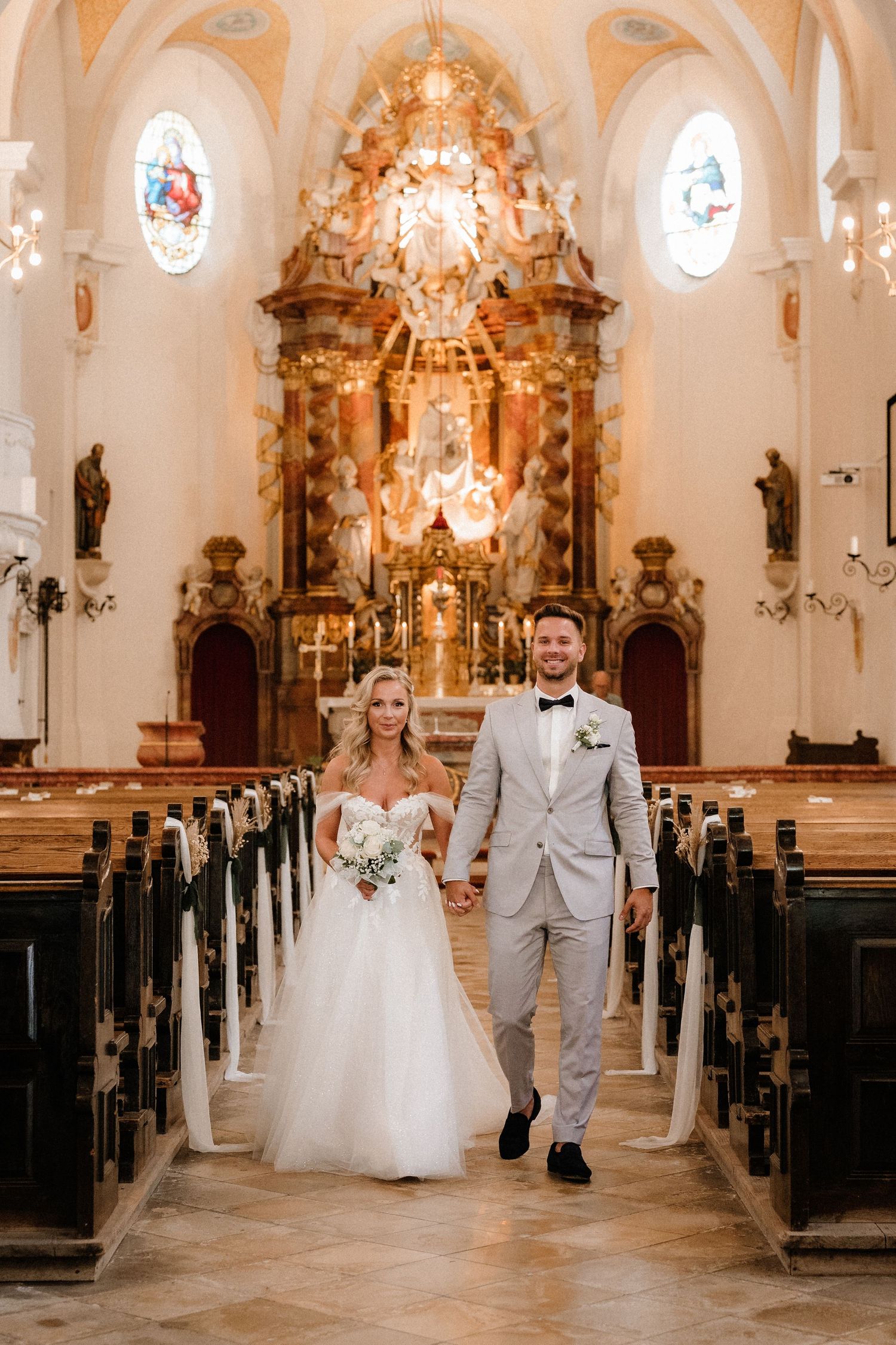 Couple walking down the aisle of an ornate Catholic church with golden altar and chandeliers.