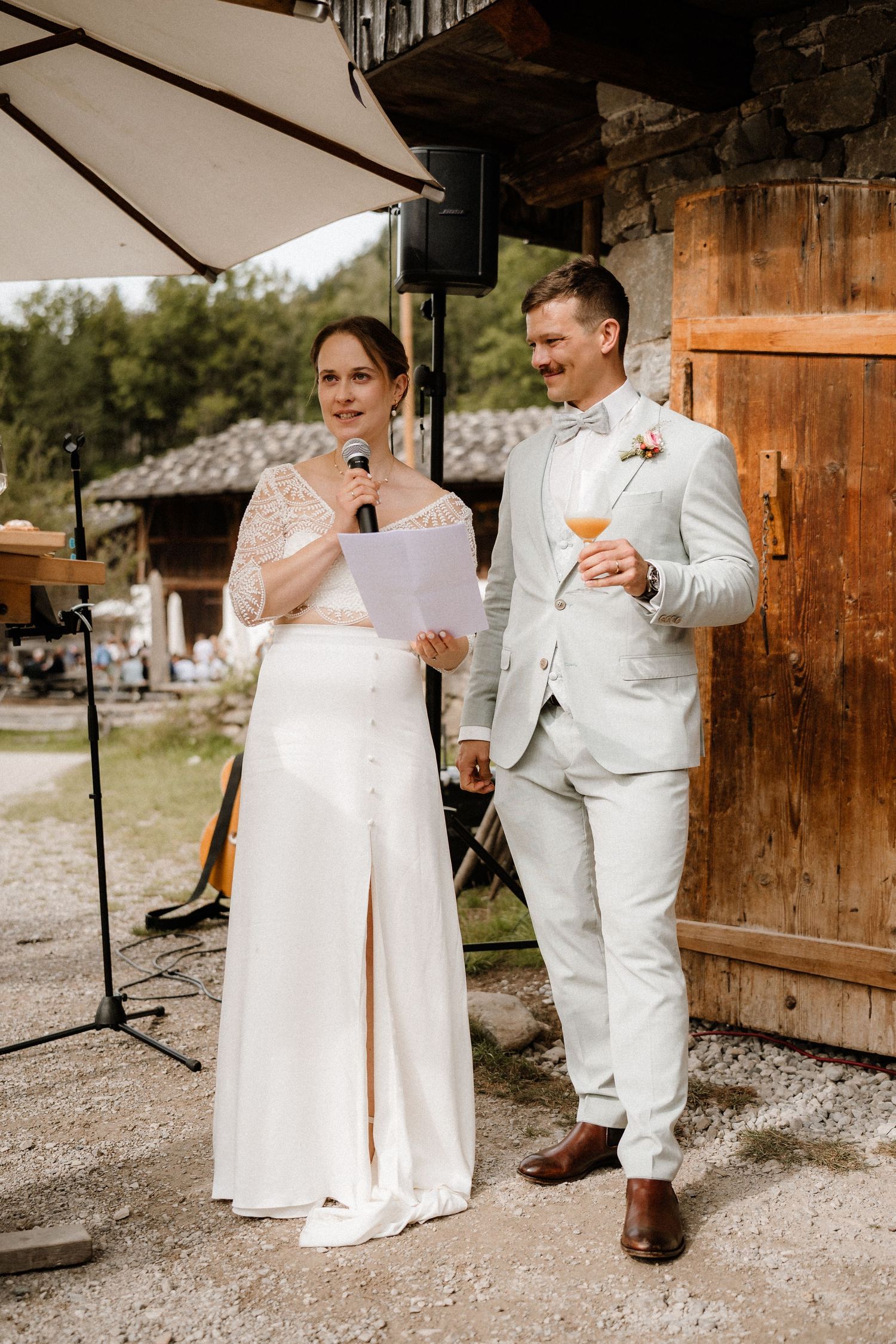 Couple in white wedding attire giving speeches under an outdoor umbrella.