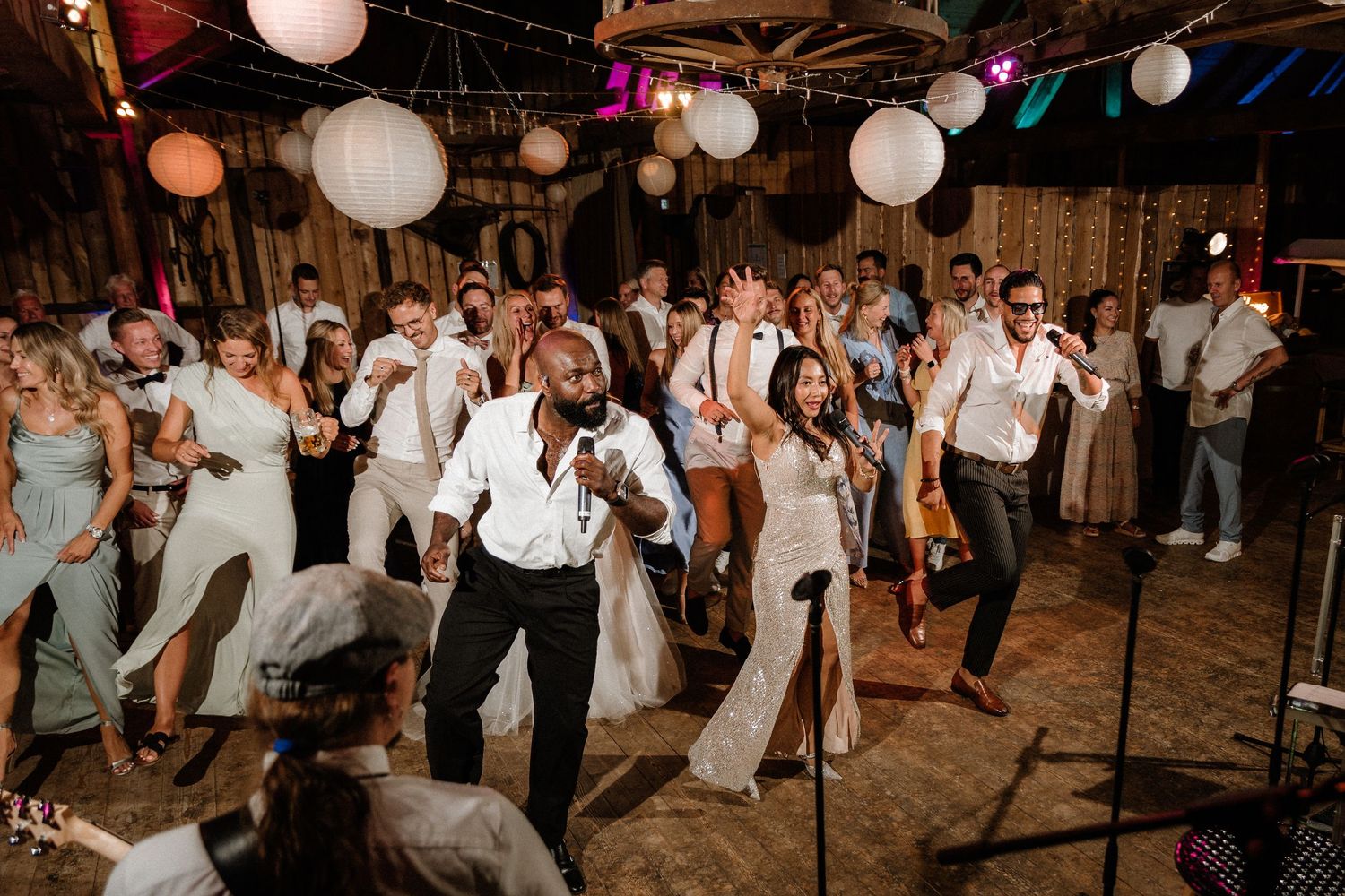 Lively dance party at a barn wedding reception with white paper lanterns overhead.