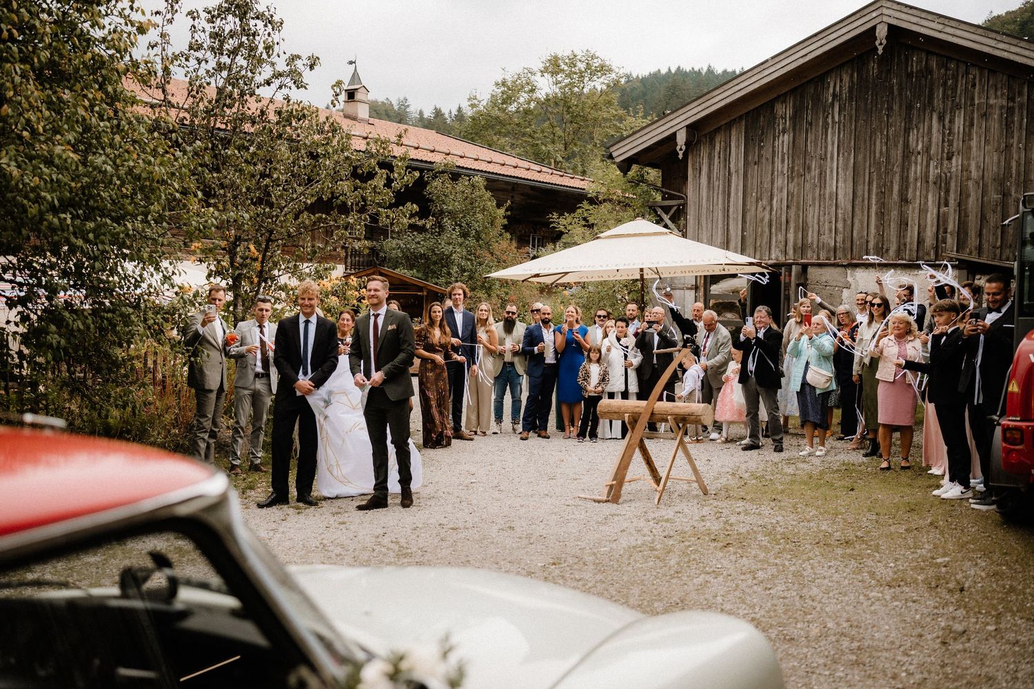 Wedding ceremony guests gather outside a rustic barn venue with a vintage car in view.