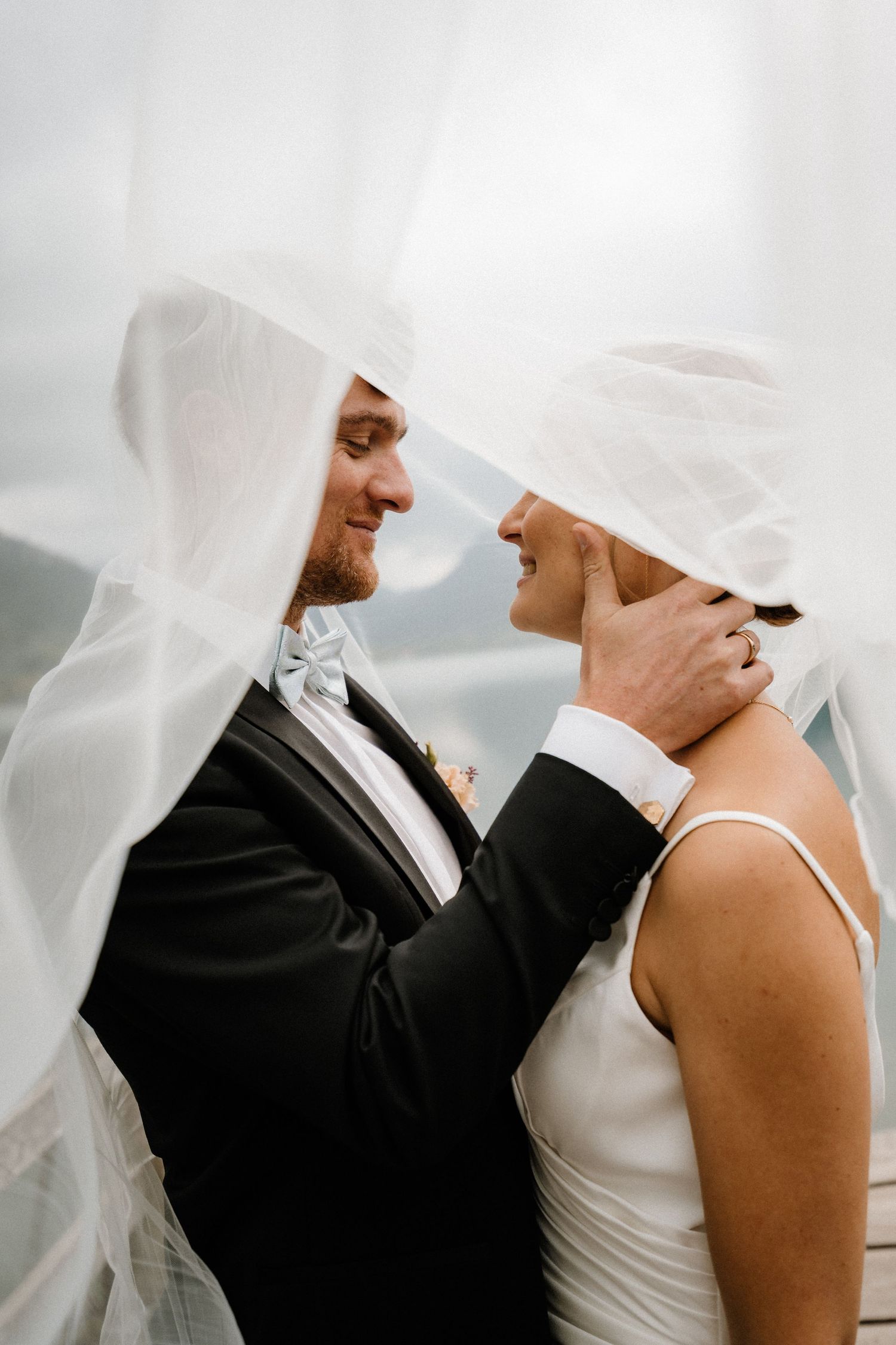 A couple shares a tender moment under a sheer white veil against a bright background.