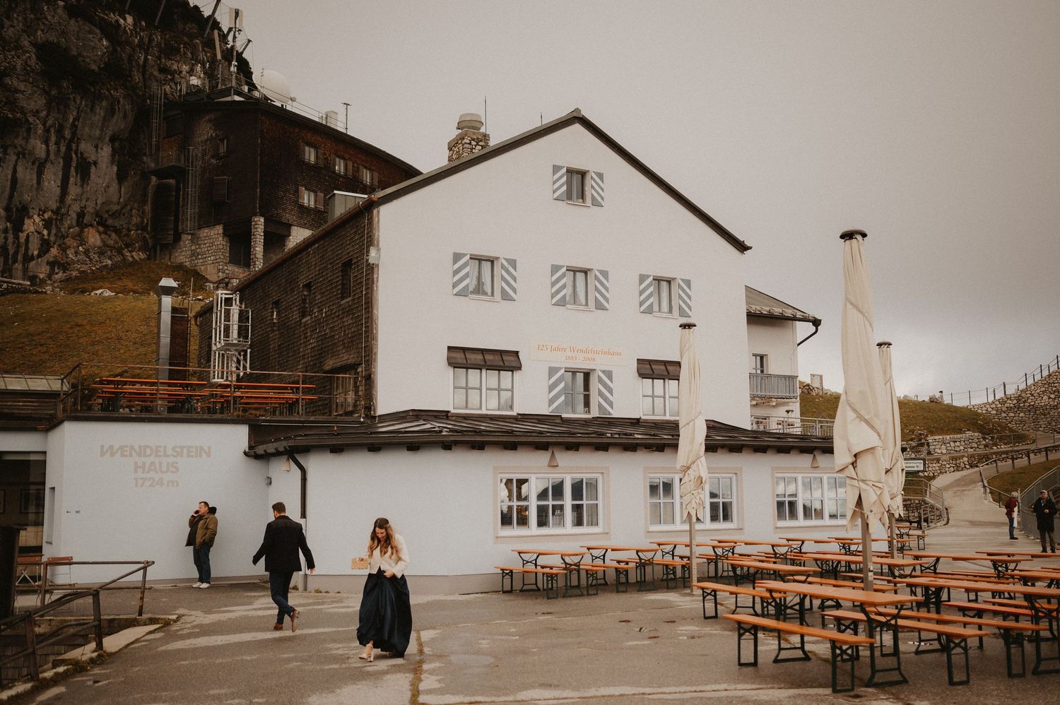 Traditional Alpine white house with outdoor seating area nestled against mountain cliffs.
