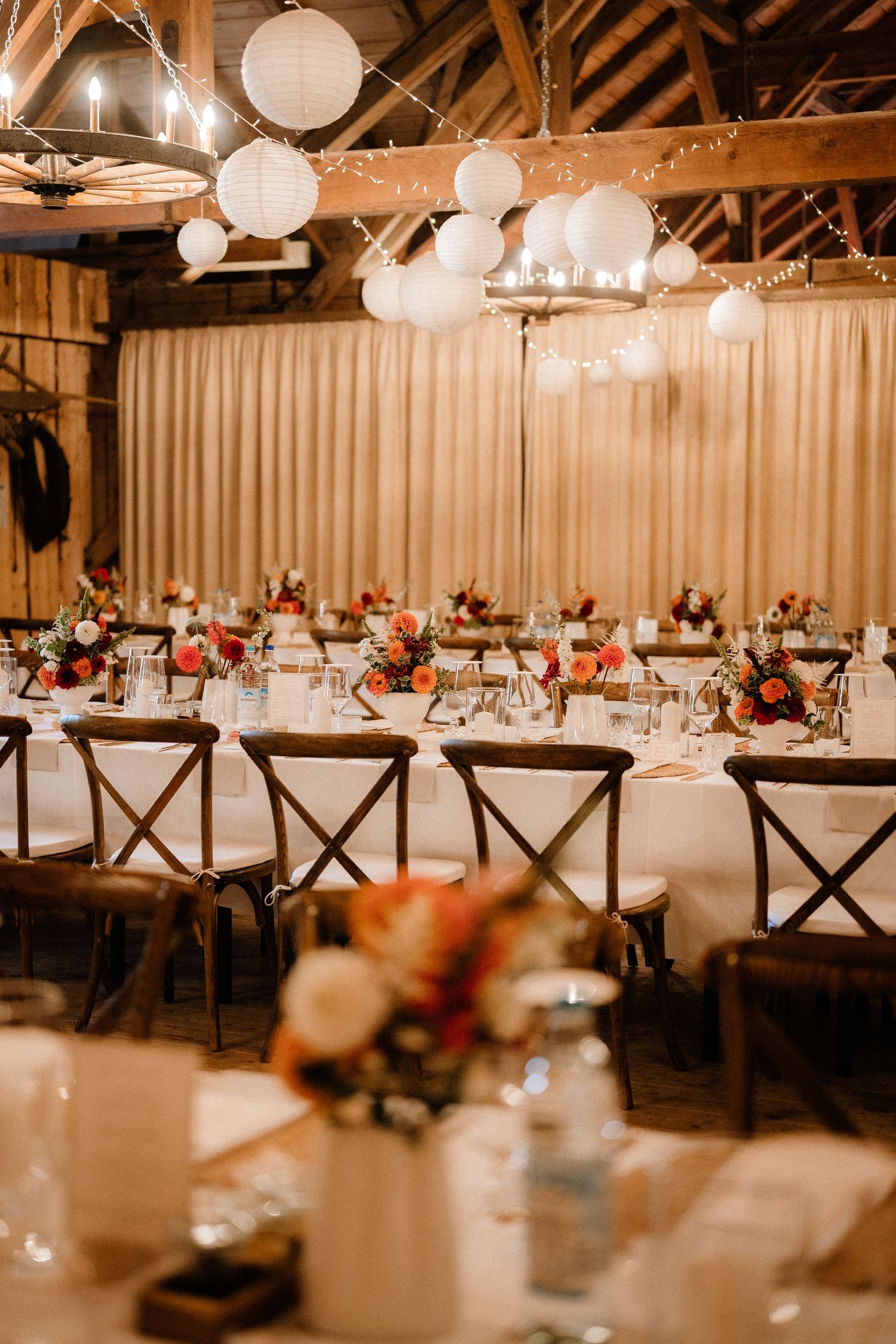 Rustic barn wedding reception venue with elegant table settings, wooden cross-back chairs, and hanging paper lanterns.