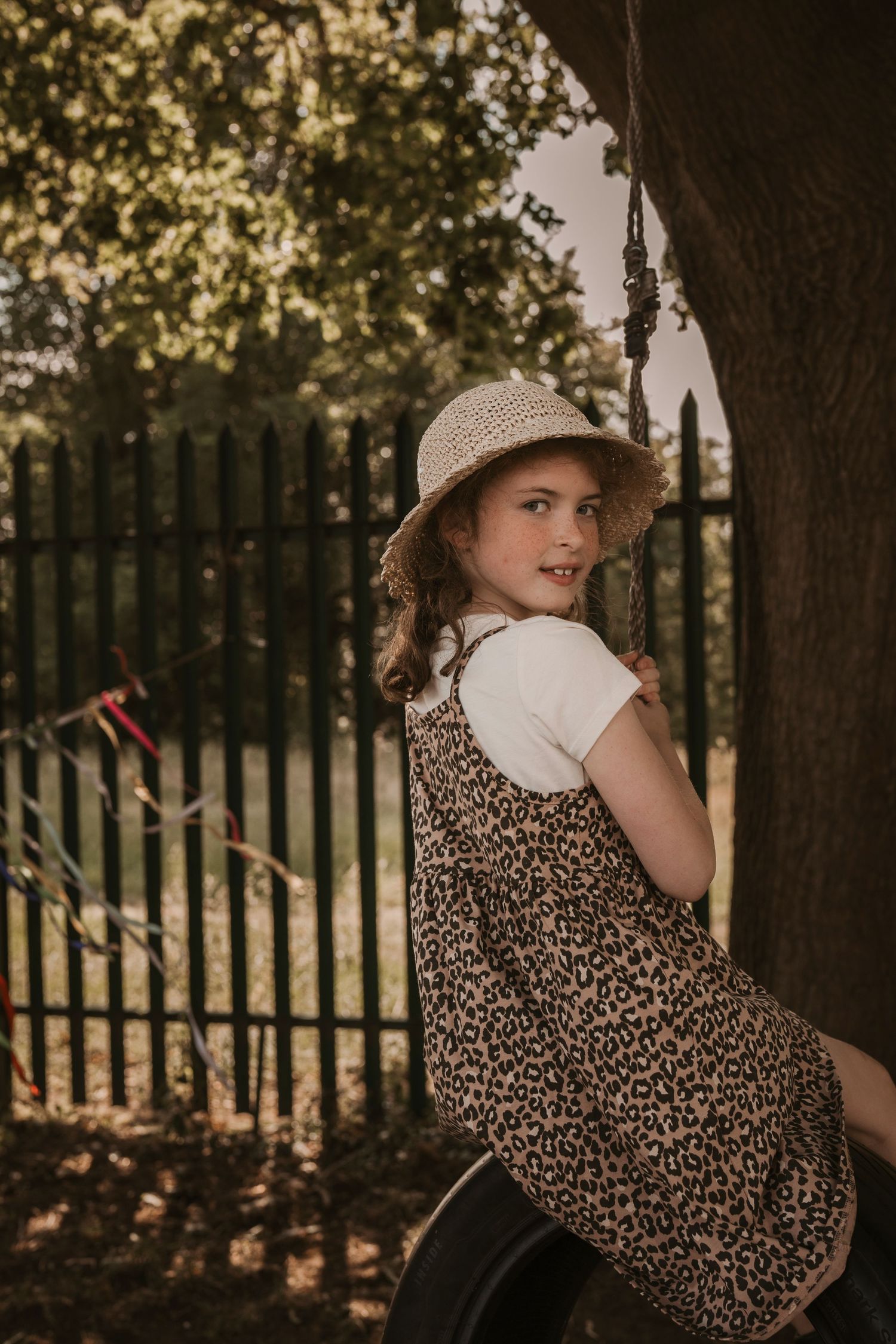 Smiling girl in a leopard-print dress and straw hat sits on a tyre swing at Roam Wild Forest School, surrounded by warm afternoon light and