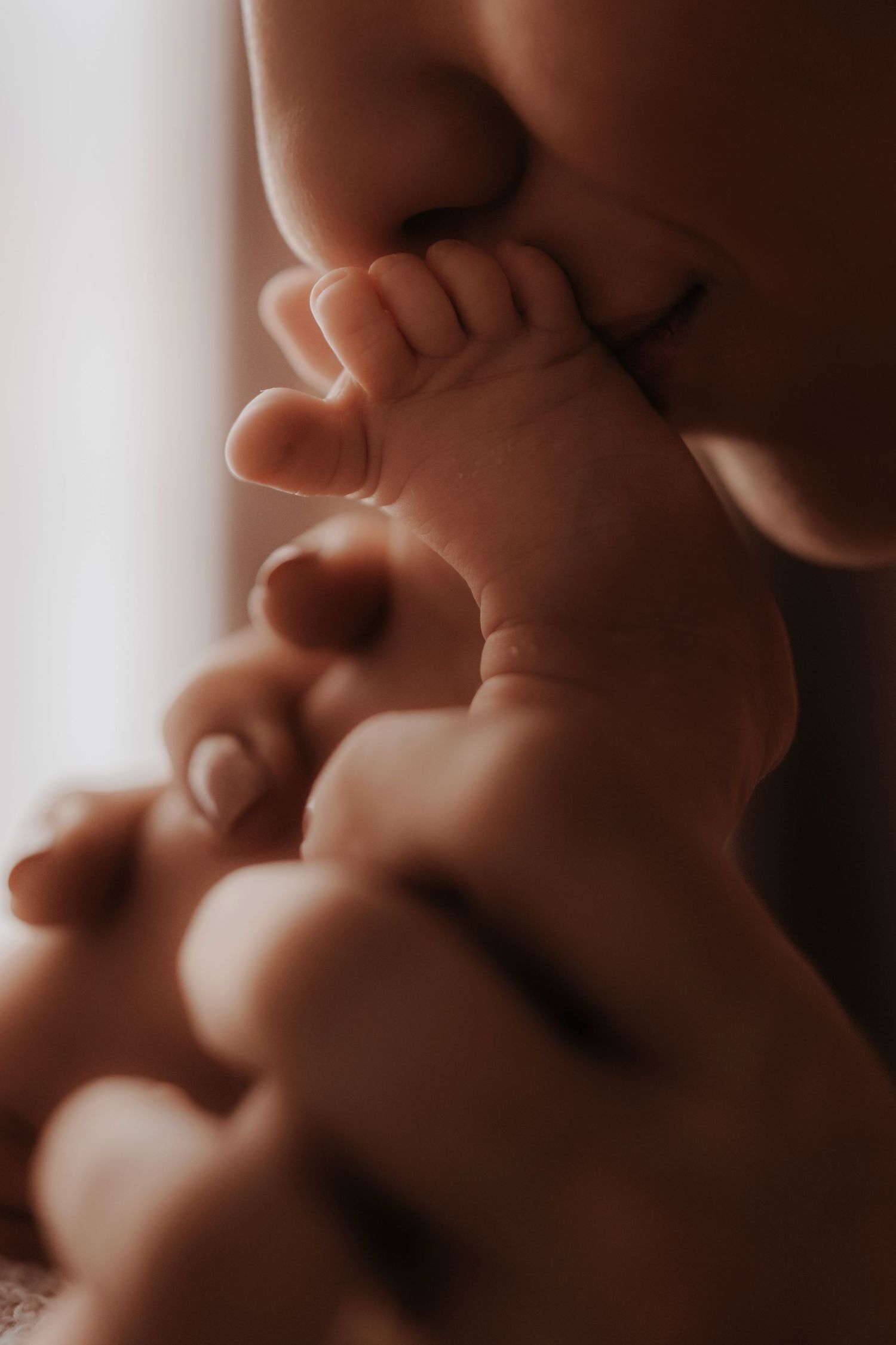 Parent kissing tiny baby feet during an intimate storytelling newborn photoshoot in soft window light.