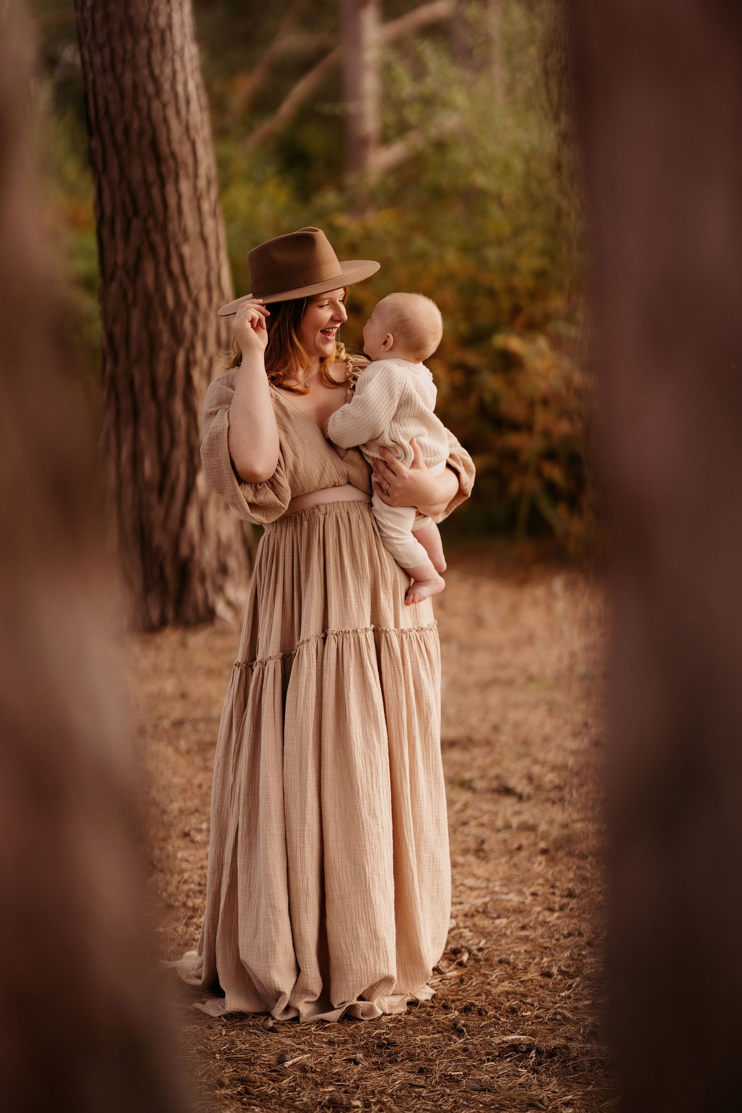 Mother standing in woodland holding her baby, wearing a hat, surrounded by trees during a calm outdoor photoshoot.