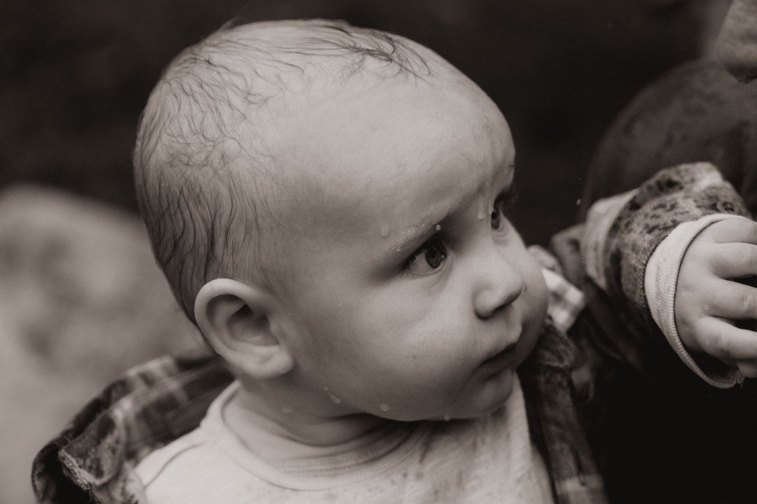 Baby with raindrops on his face gazes curiously outdoors, captured in timeless black and white photography.