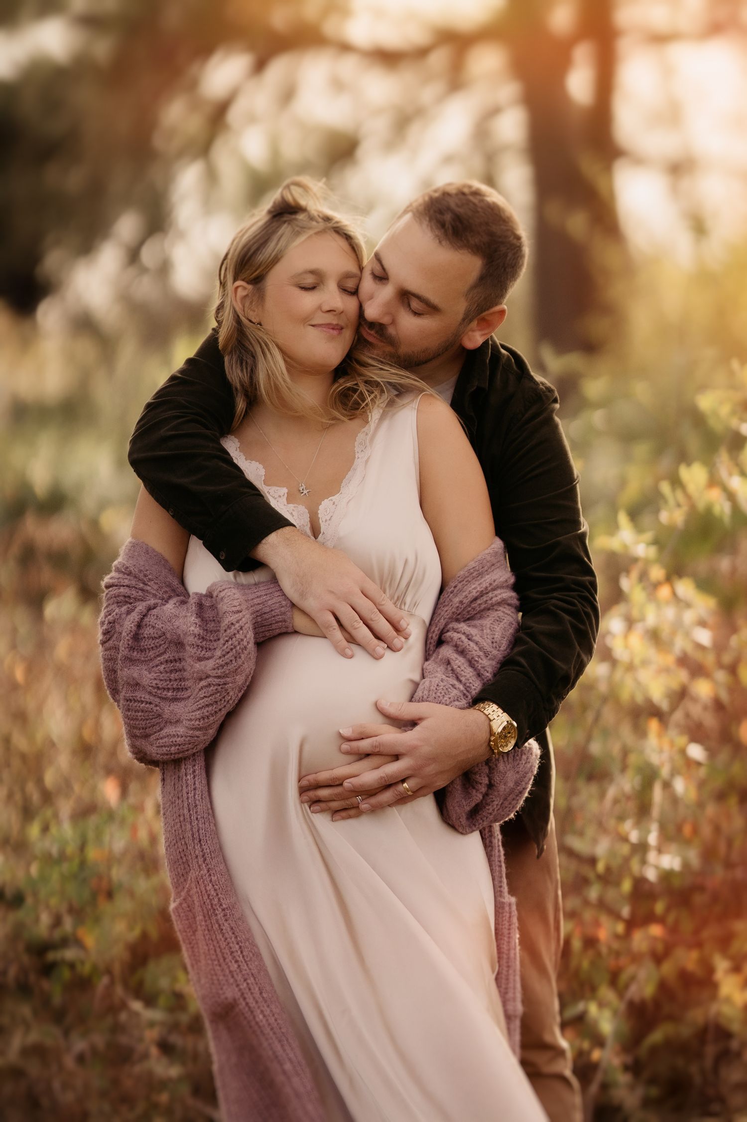 Expecting couple embracing outdoors in golden hour light, holding a pregnant belly during a maternity photography session.