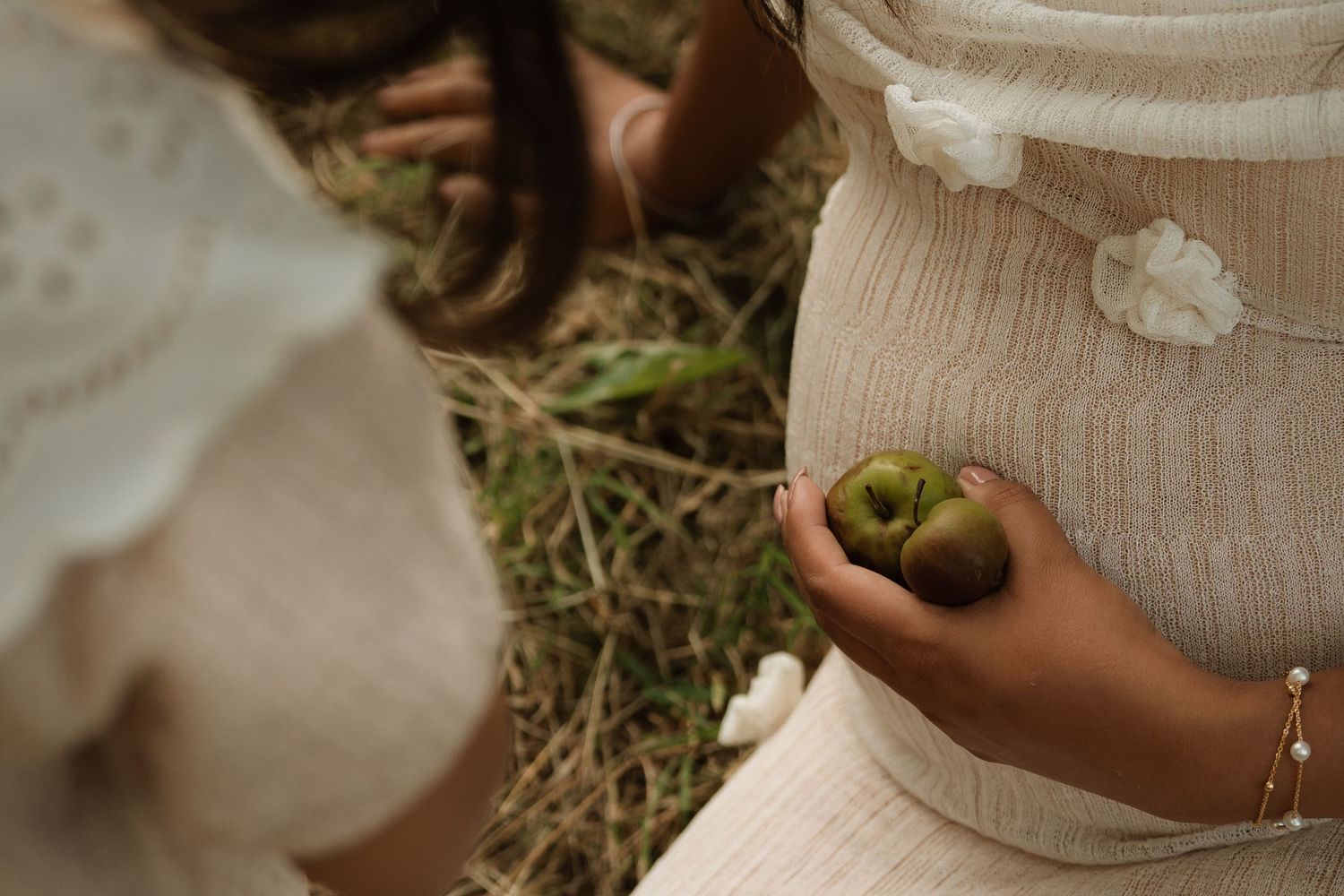 Little girl places a green apple on her mother’s baby bump during an outdoor maternity session.