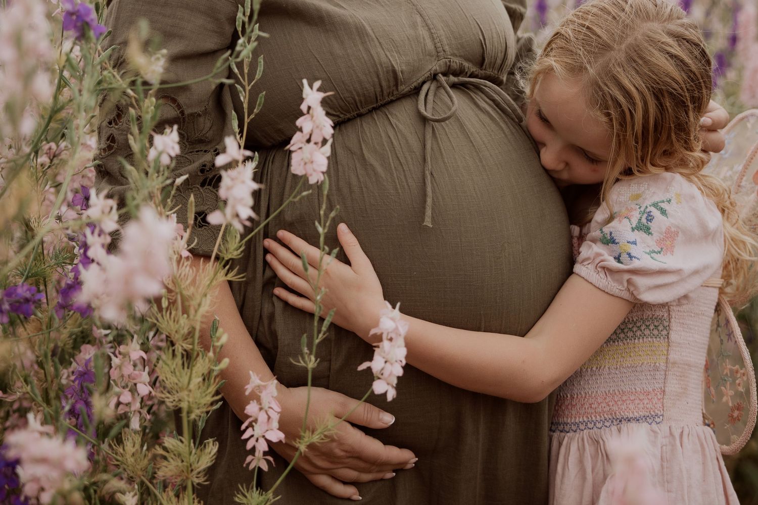 A little girl hugging her mother’s pregnant belly among blooming delphiniums – tender maternity session in the Confetti Flower Fields near B