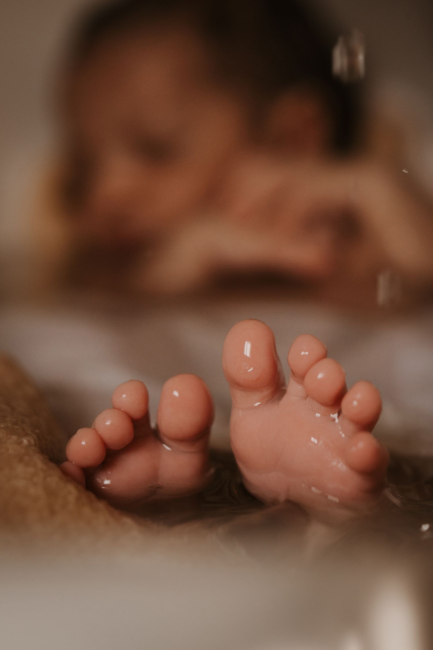 Newborn’s tiny feet resting in warm bath water, softly lit, with the baby’s face blurred in the background during a quiet bathing moment