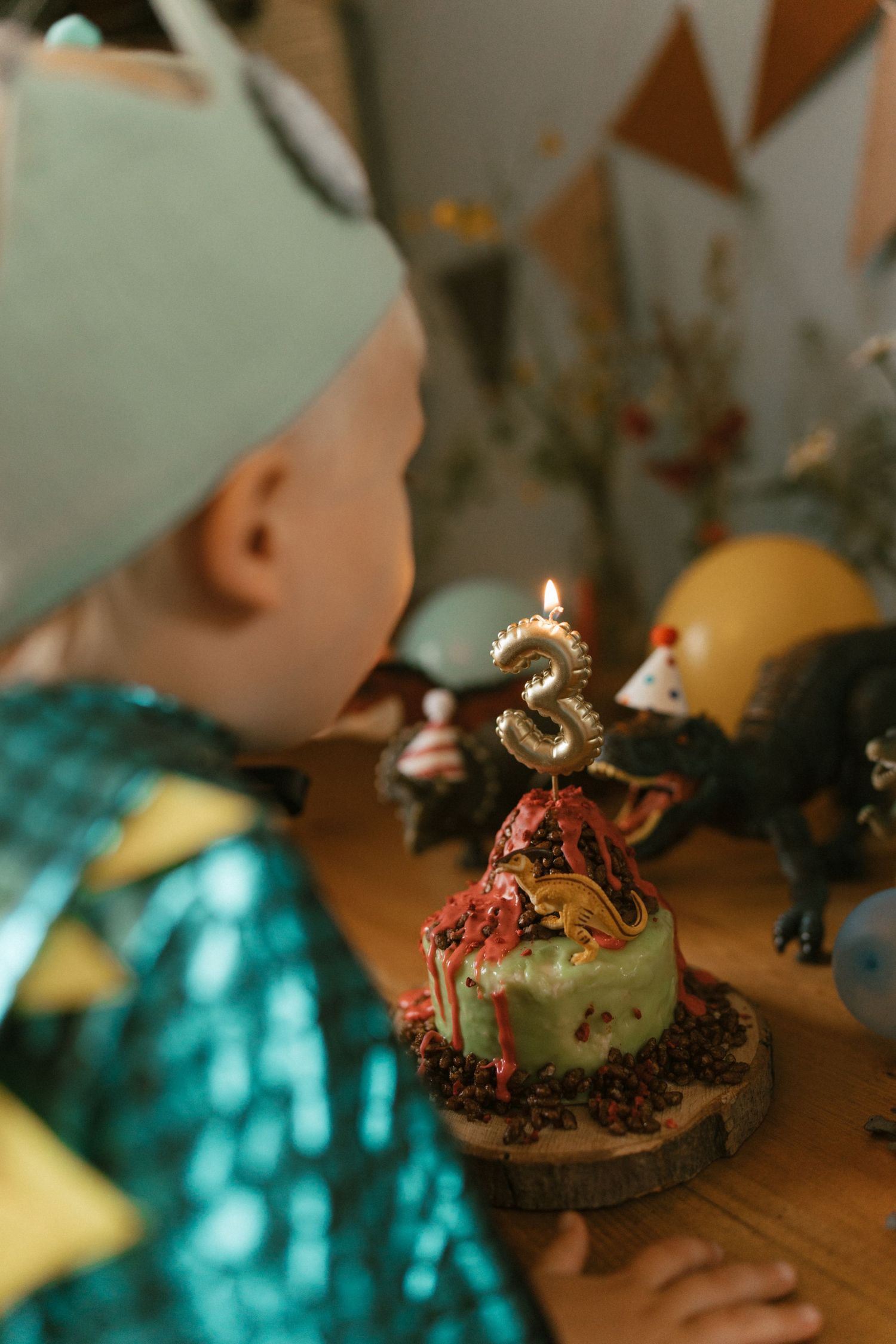 Toddler wearing a crown and party outfit leaning toward a small birthday cake with a lit candle shaped like the number one, surrounded by so
