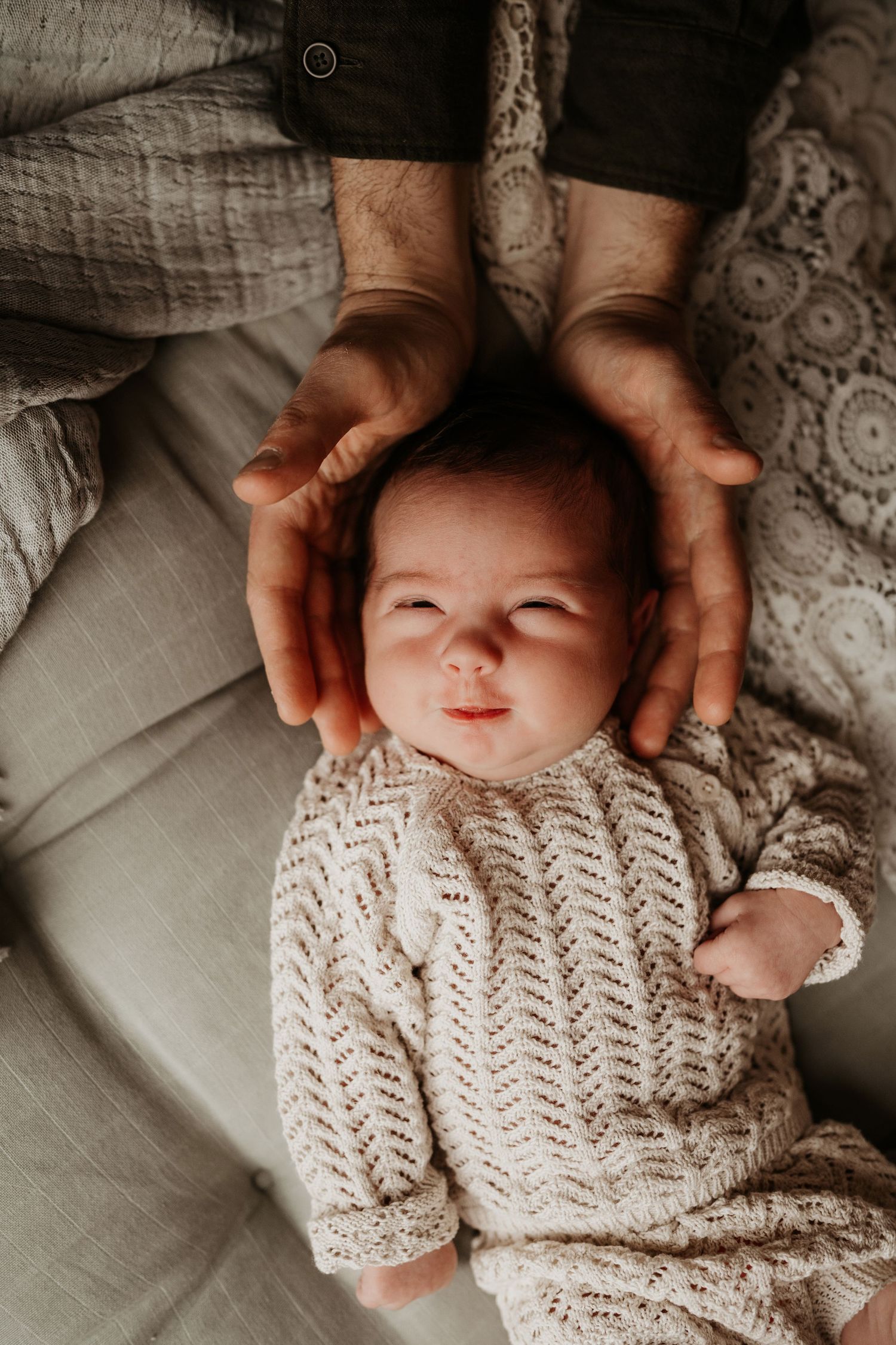 Newborn lying on a soft bed with parents’ hands framing their head, a tender moment captured in a storytelling newborn photoshoot by Angela