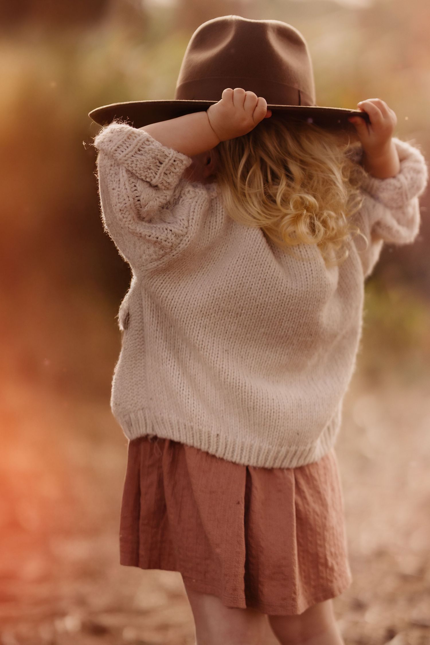 Toddler seen from behind adjusting a hat, standing in warm autumn light during a quiet outdoor photoshoot.