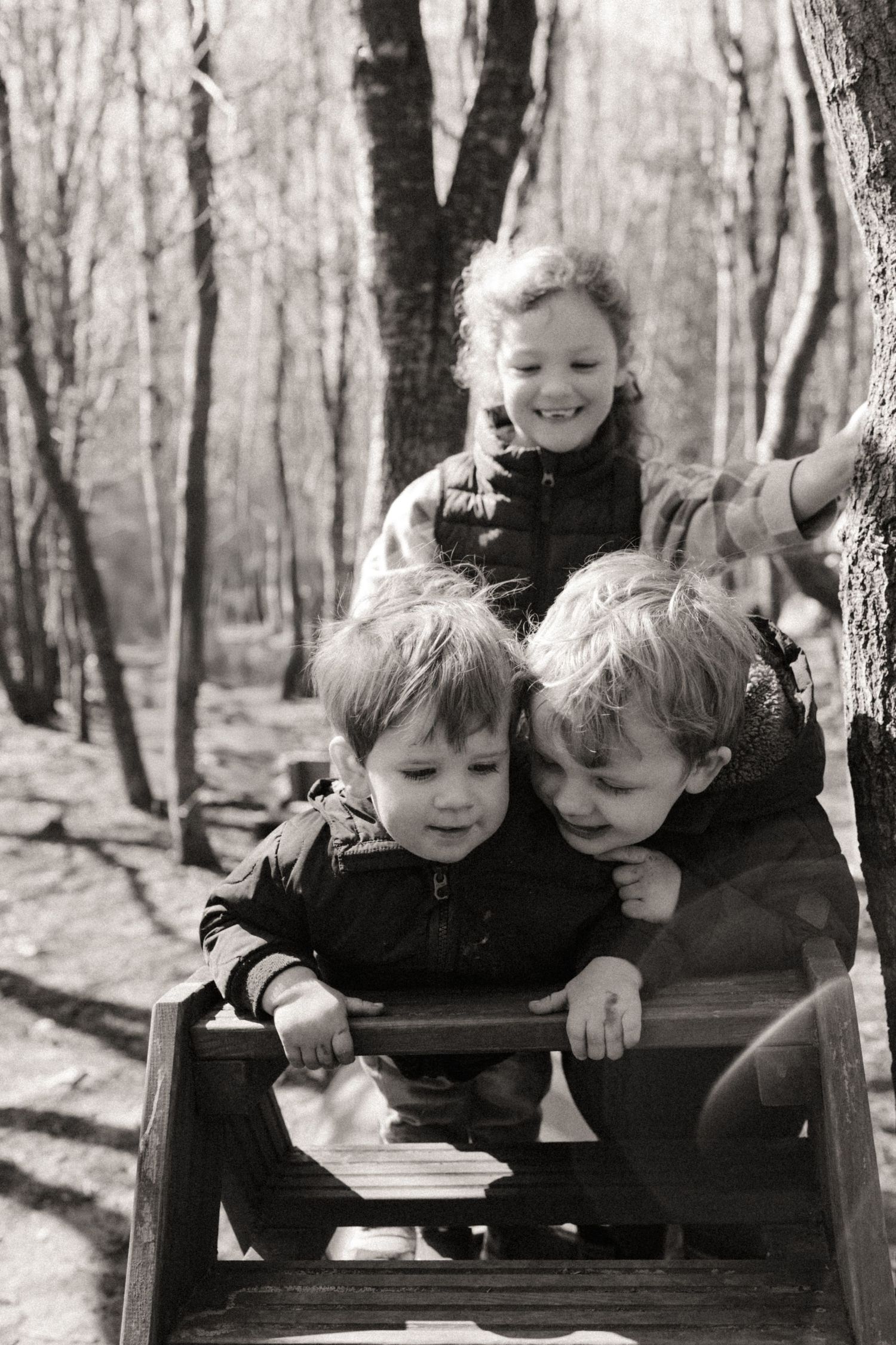 Three siblings play and laugh together in the woods during a natural childhood photography session in Little Wildlings