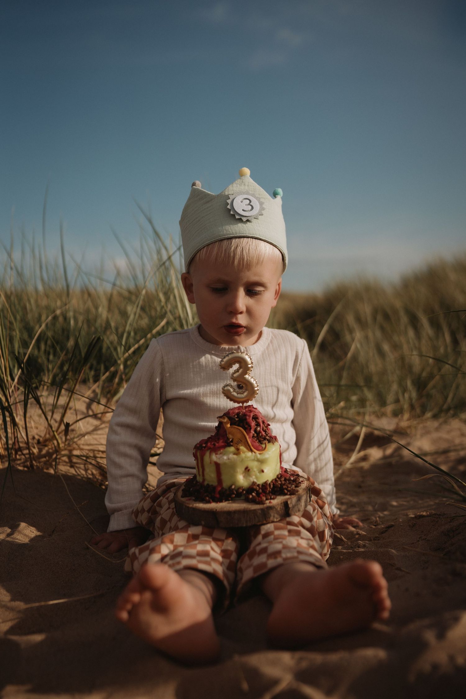 Toddler wearing a crown sits on the beach with a birthday cake during a seaside family photoshoot.