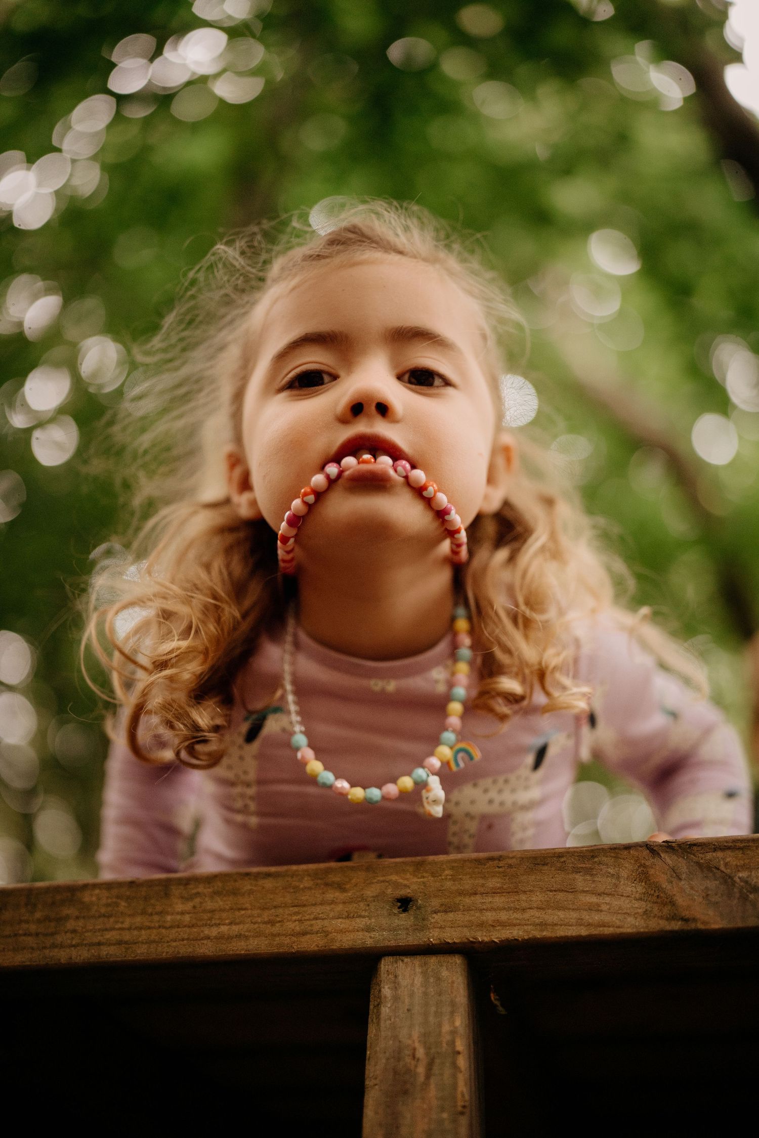 Little girl peeks over a wooden ledge, sunlight and beads catching the soft forest light during Little Wildlings Photoshoot