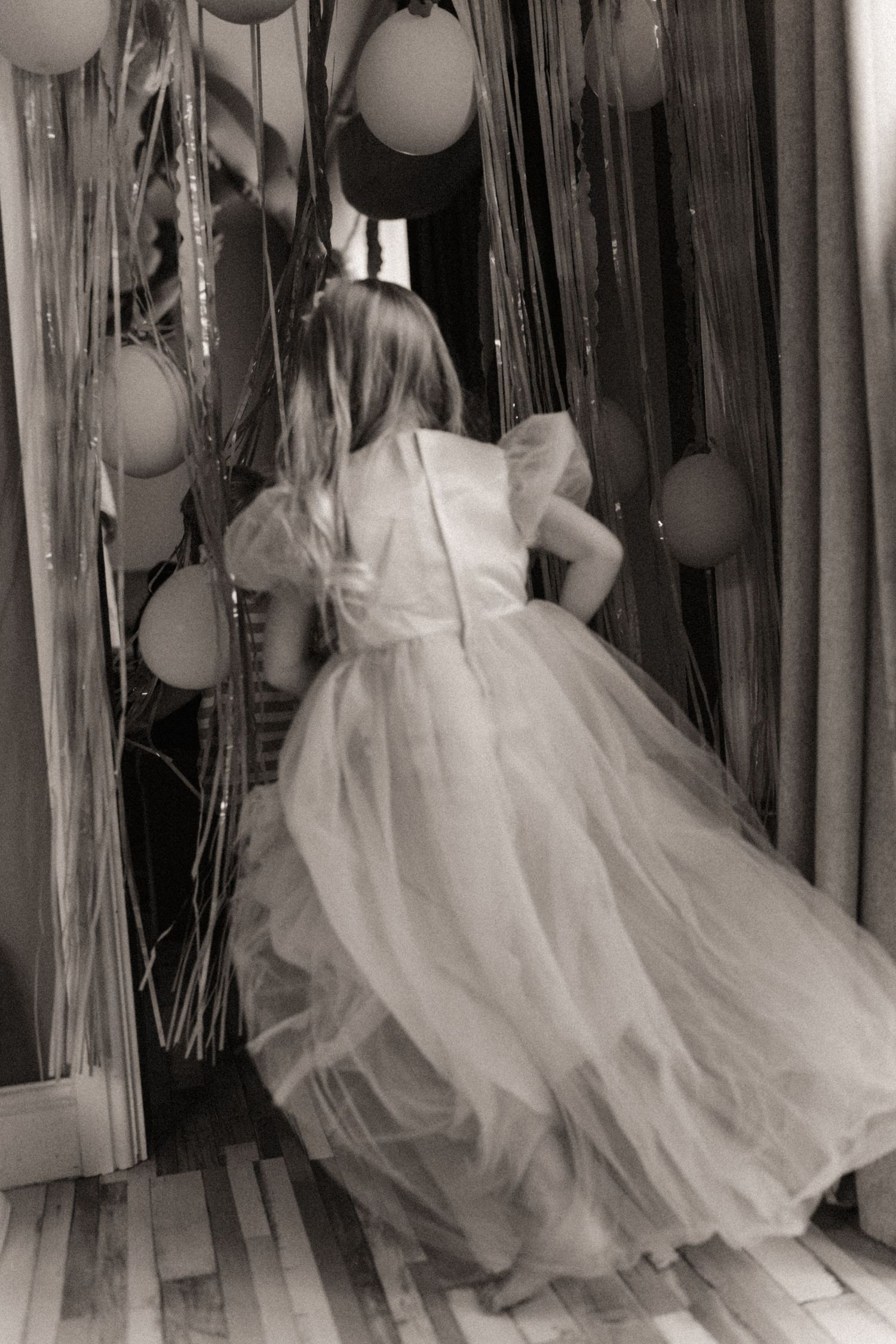 Black and white image of a girl in a flowing dress stepping into a room filled with balloons and streamers, seen from behind during a playfu