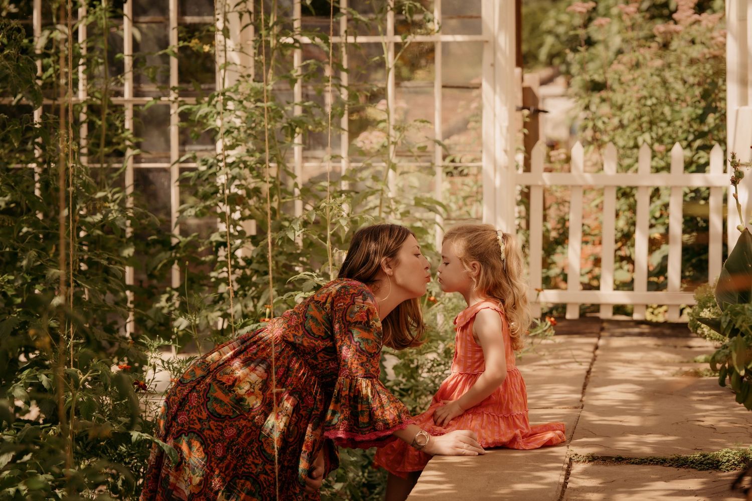 Parent kneeling to kiss a young child in a sunlit garden during a family session.