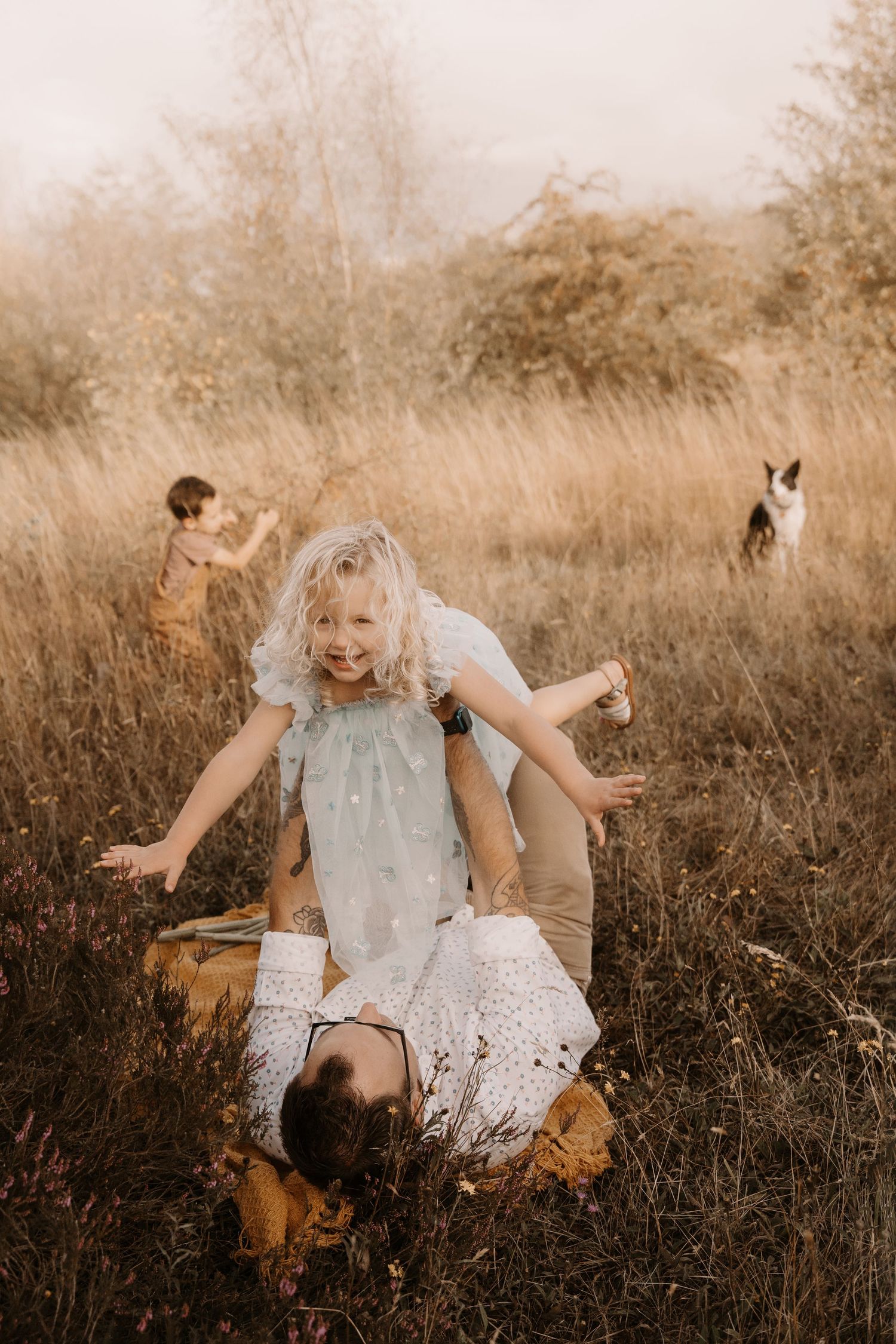 Girl plays with dad in a golden field with her dog during a joyful outdoor family photoshoot in Swadlincote