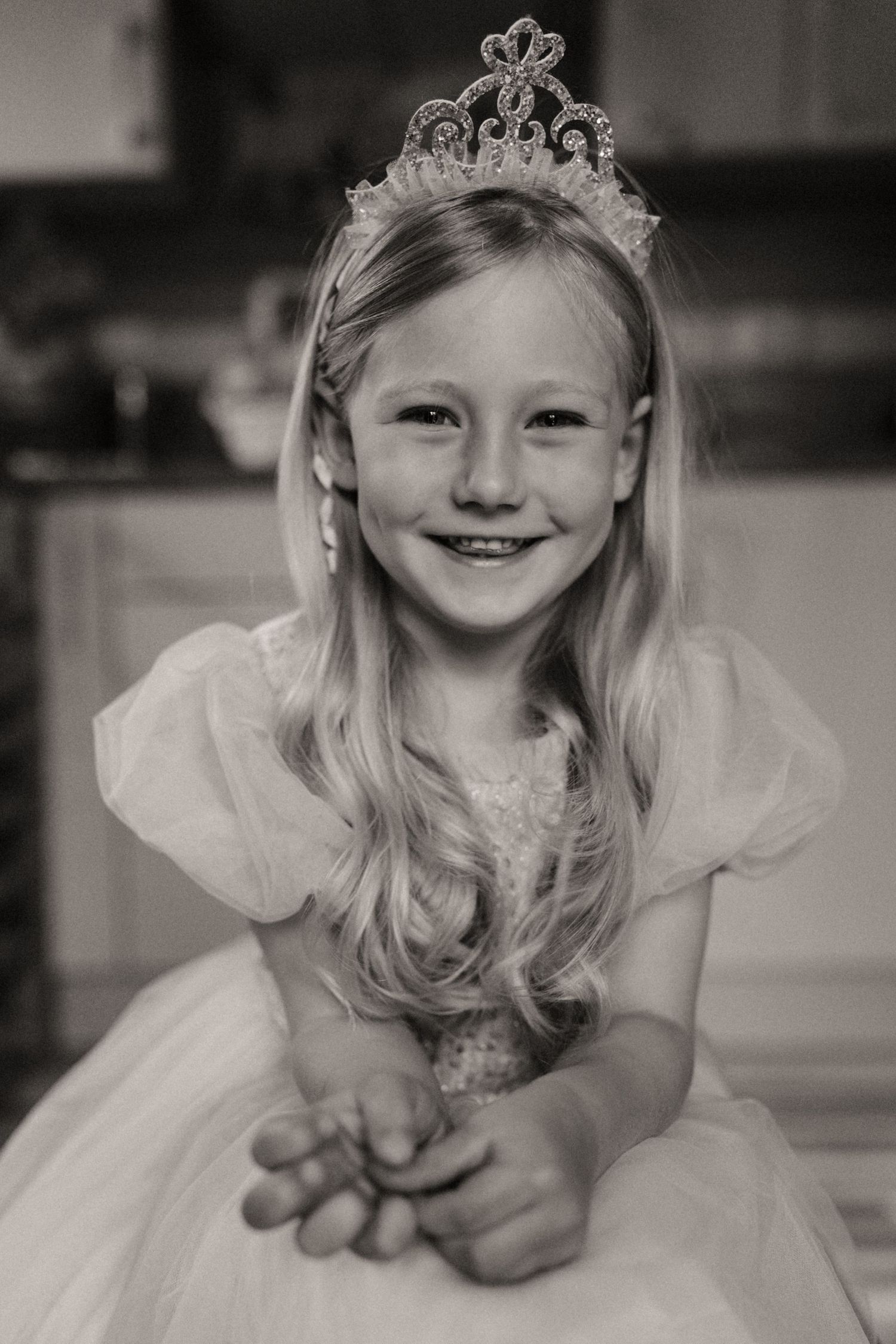 Black and white portrait of a young girl wearing a tiara and tulle dress, smiling shyly while sitting on the floor during a childhood photos