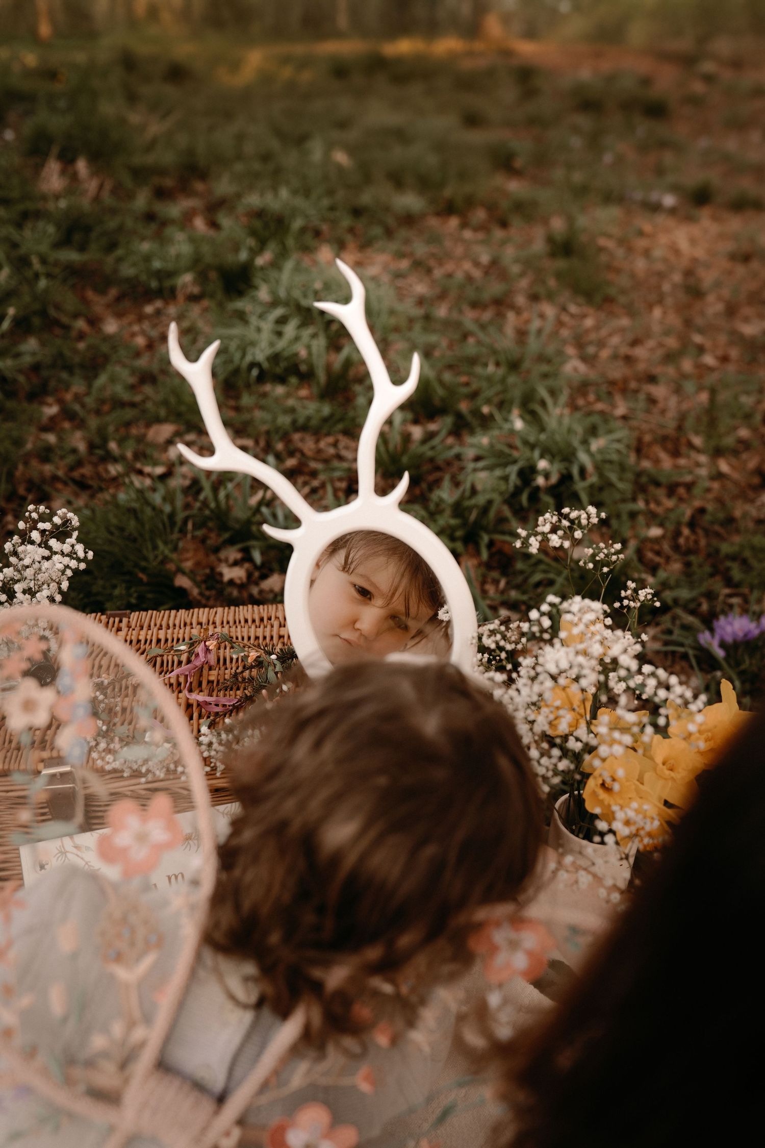 Child looking at her reflection in a mirror with whimsical antlers surrounded by wildflowers at a nature-inspired mother's day photoshoot