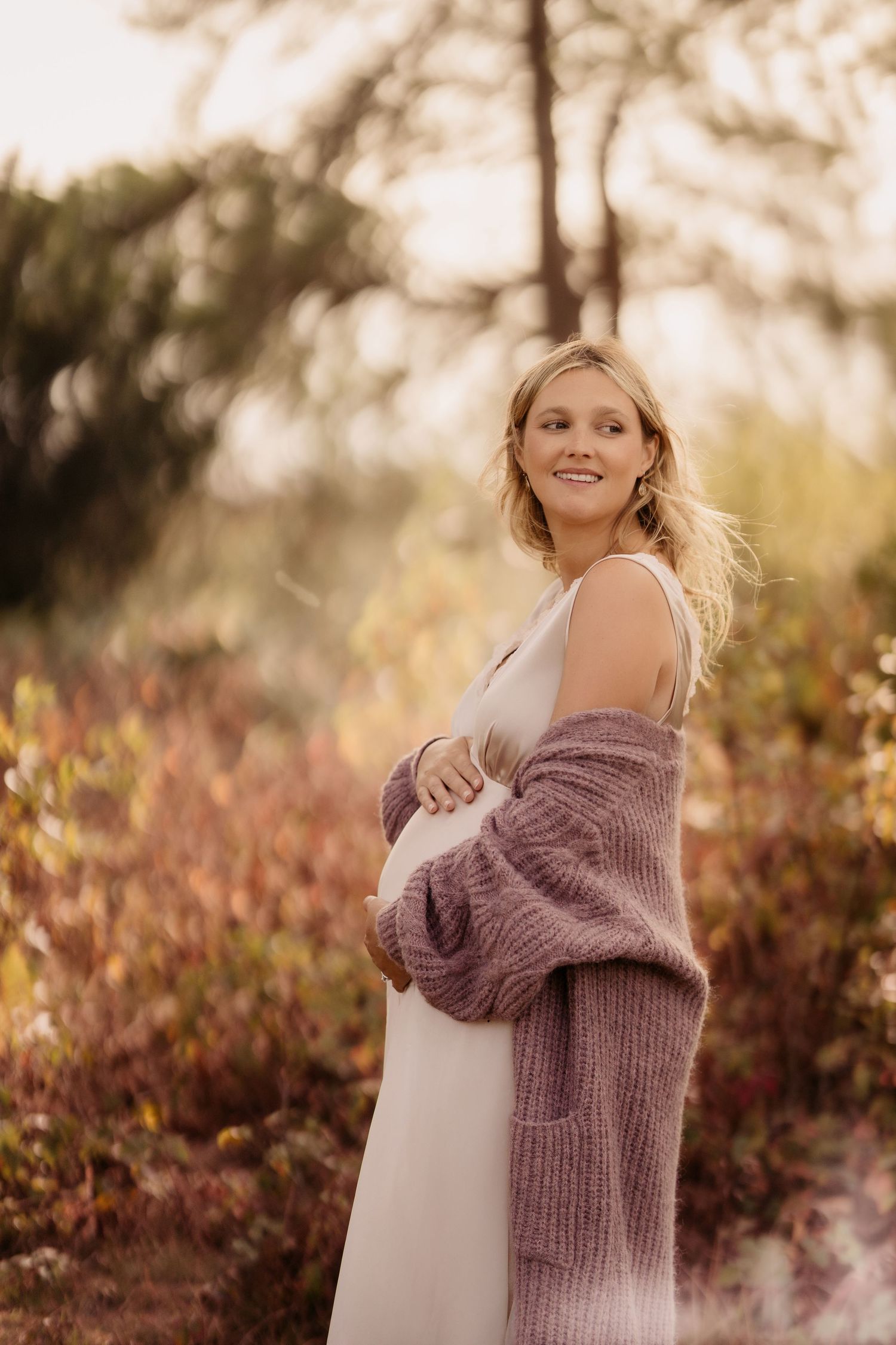 Pregnant parent standing outdoors in warm light during a relaxed maternity session.
