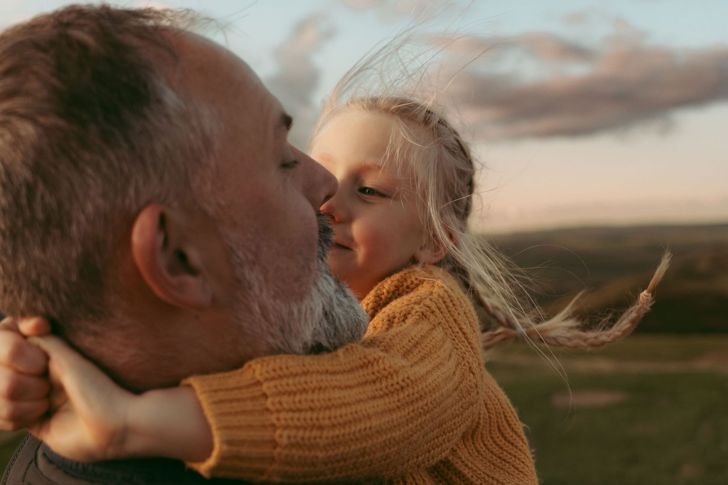Young child hugging and kissing an older man outdoors, hair blowing in the wind with an open landscape behind them
