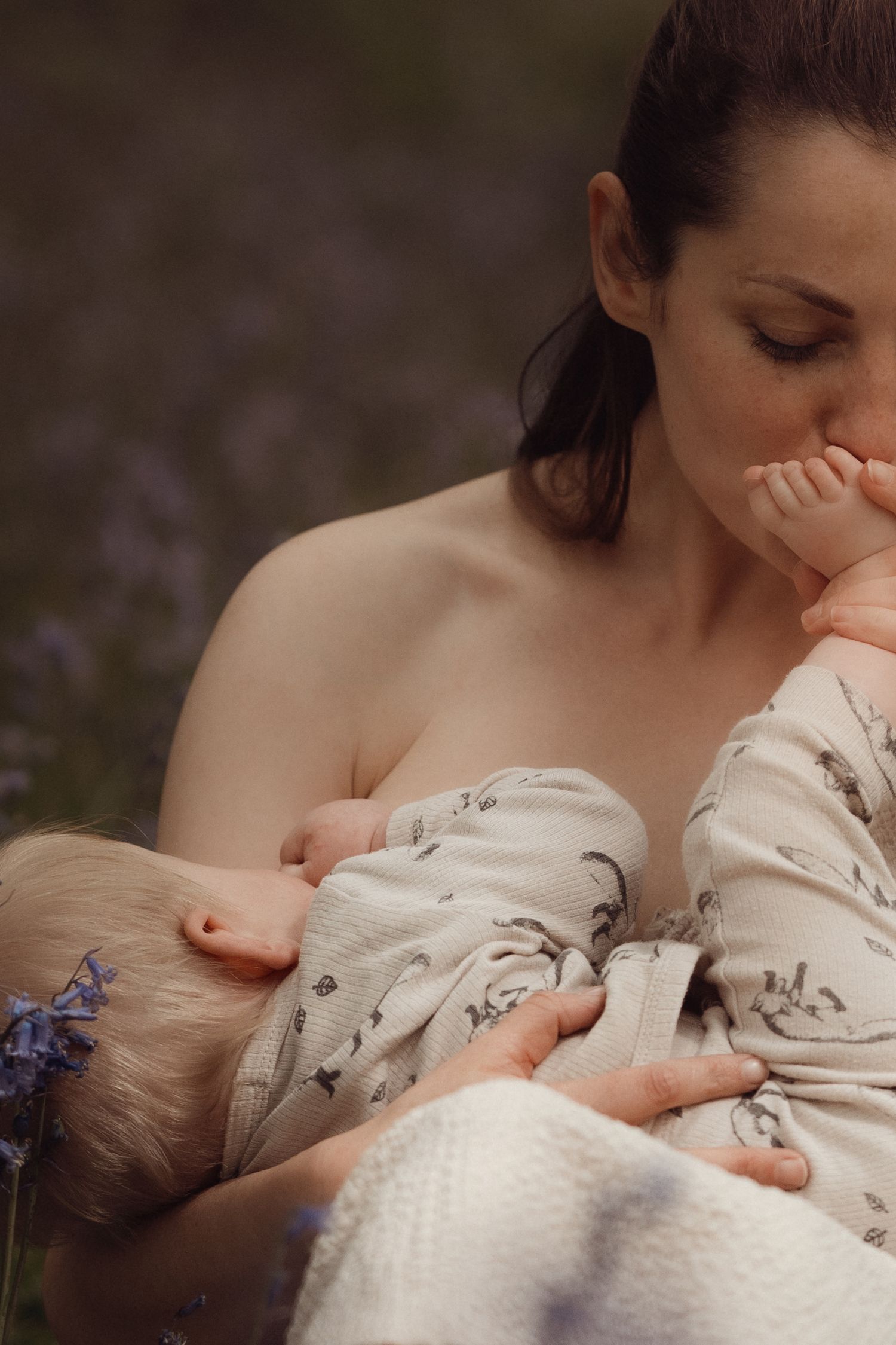 Mother breastfeeding her baby outdoors, cradling the child close to her chest, surrounded by soft purple wildflowers in gentle natural light