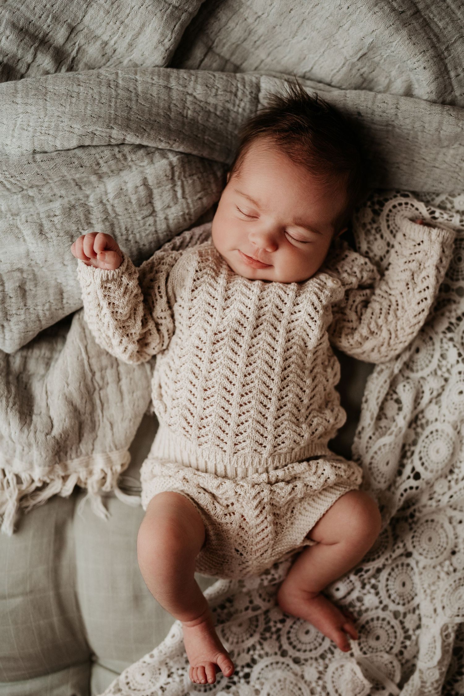 Newborn resting on a textured blanket in a knitted outfit, photographed in a warm, natural style during an in-home newborn lifestyle session