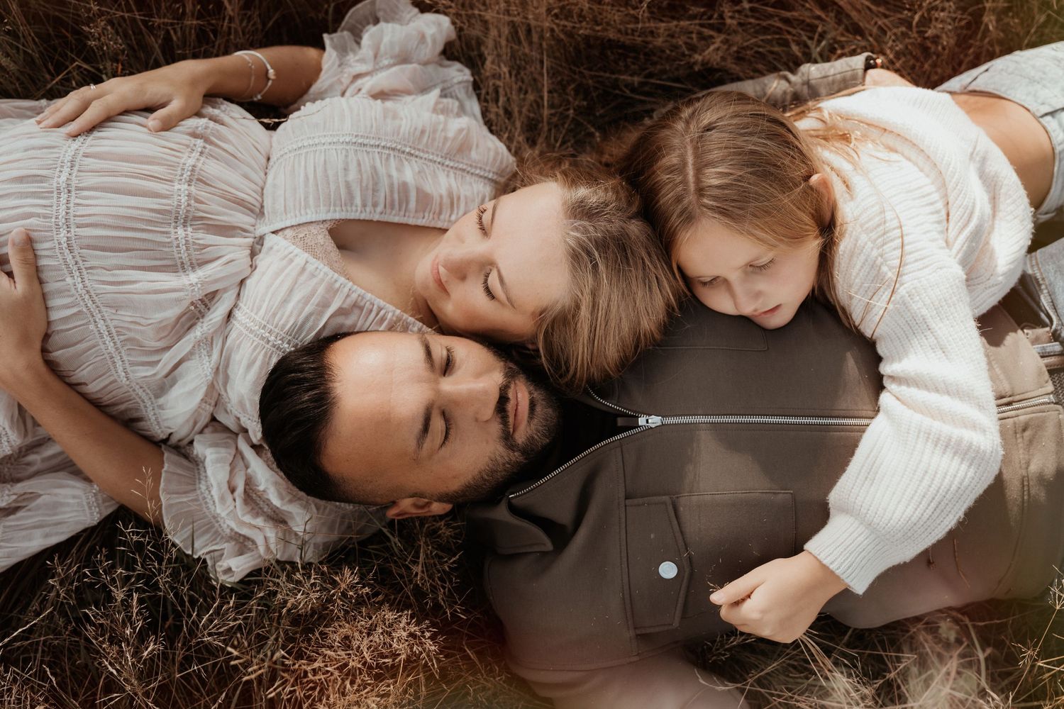 Family lies together in tall grass sharing smiles during a warm autumn Maternity photoshoot outdoors in Hugglescote
