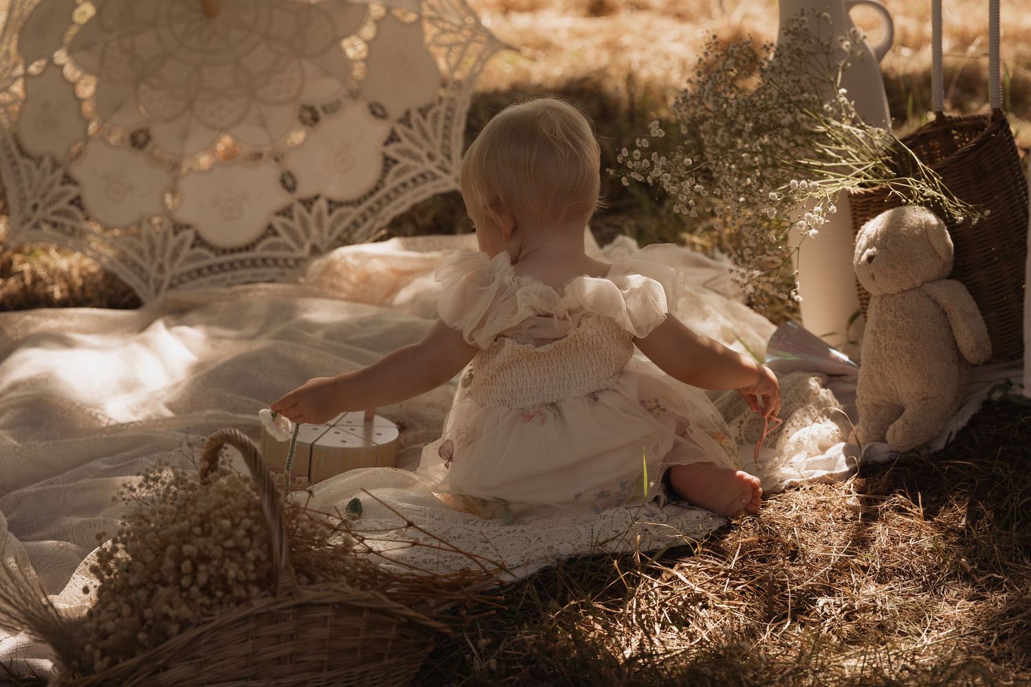 Baby sitting on a blanket surrounded by lace and toys during a gentle outdoor documentary childhood first birthday photoshoot