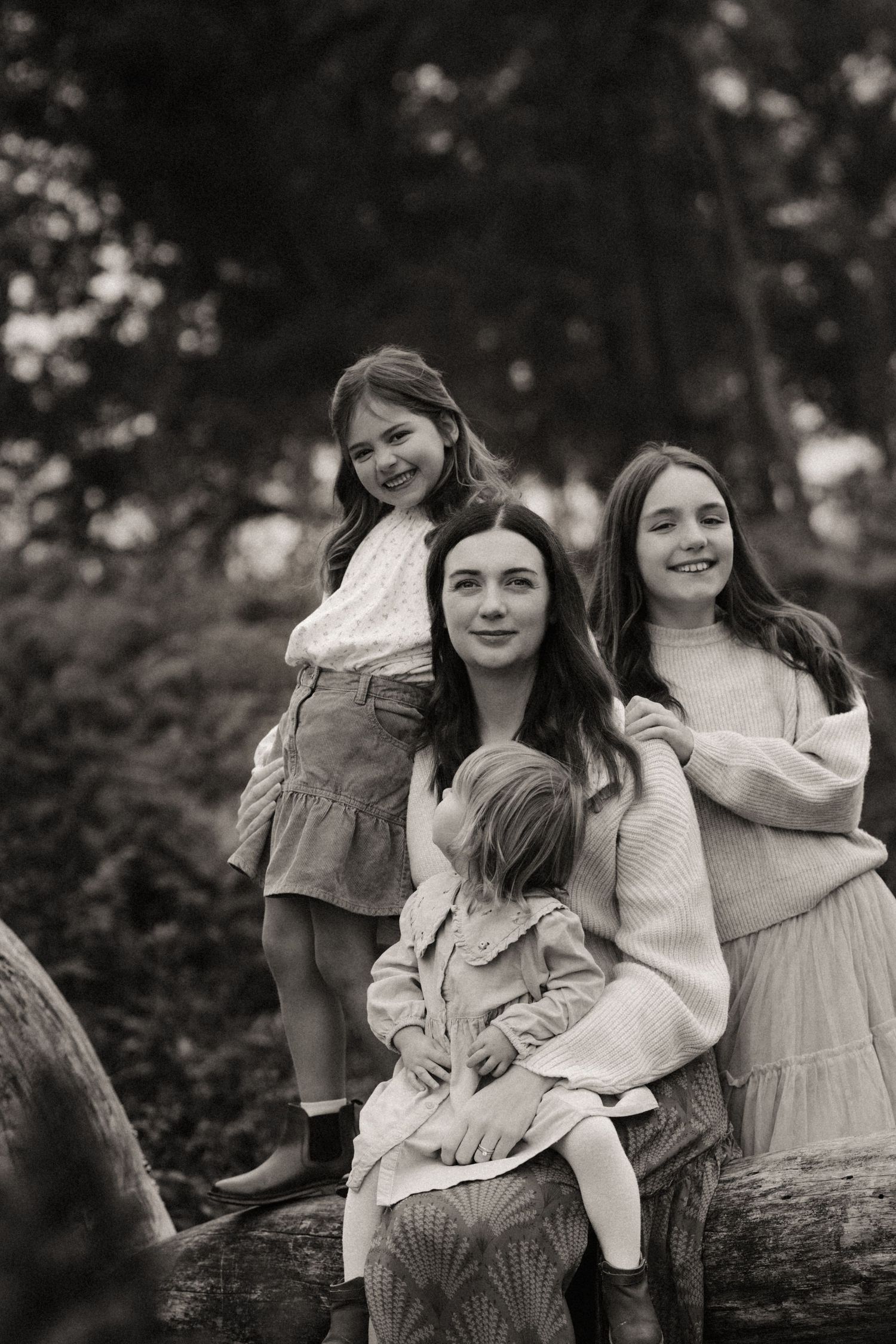 Mother and daughters smile together on a fallen log during a warm woodland family photoshoot in Birmingham, Lickey Hills