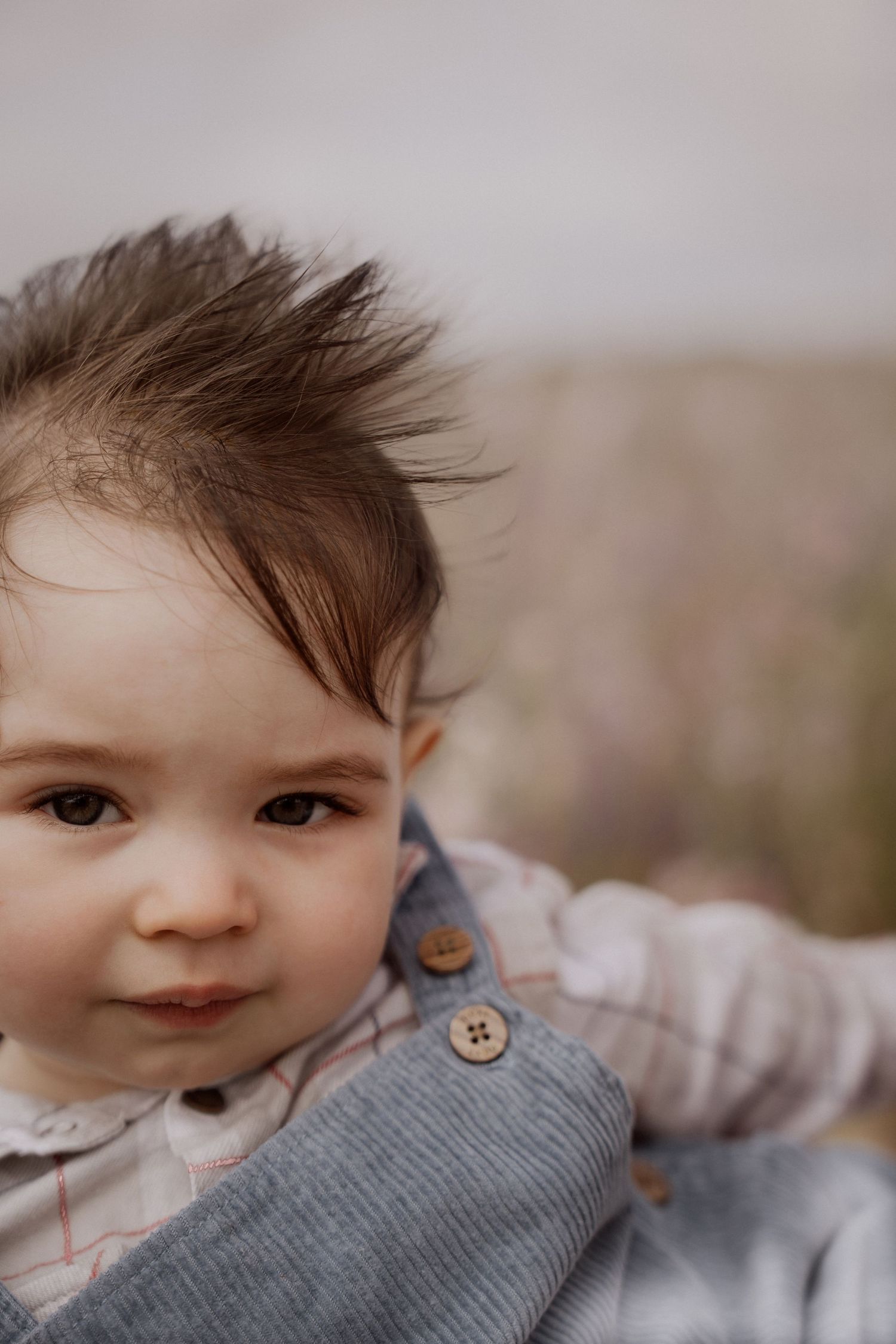 Curious baby boy with windswept hair sits among wildflowers, wearing soft overalls during the Confetti Field photoshoot
