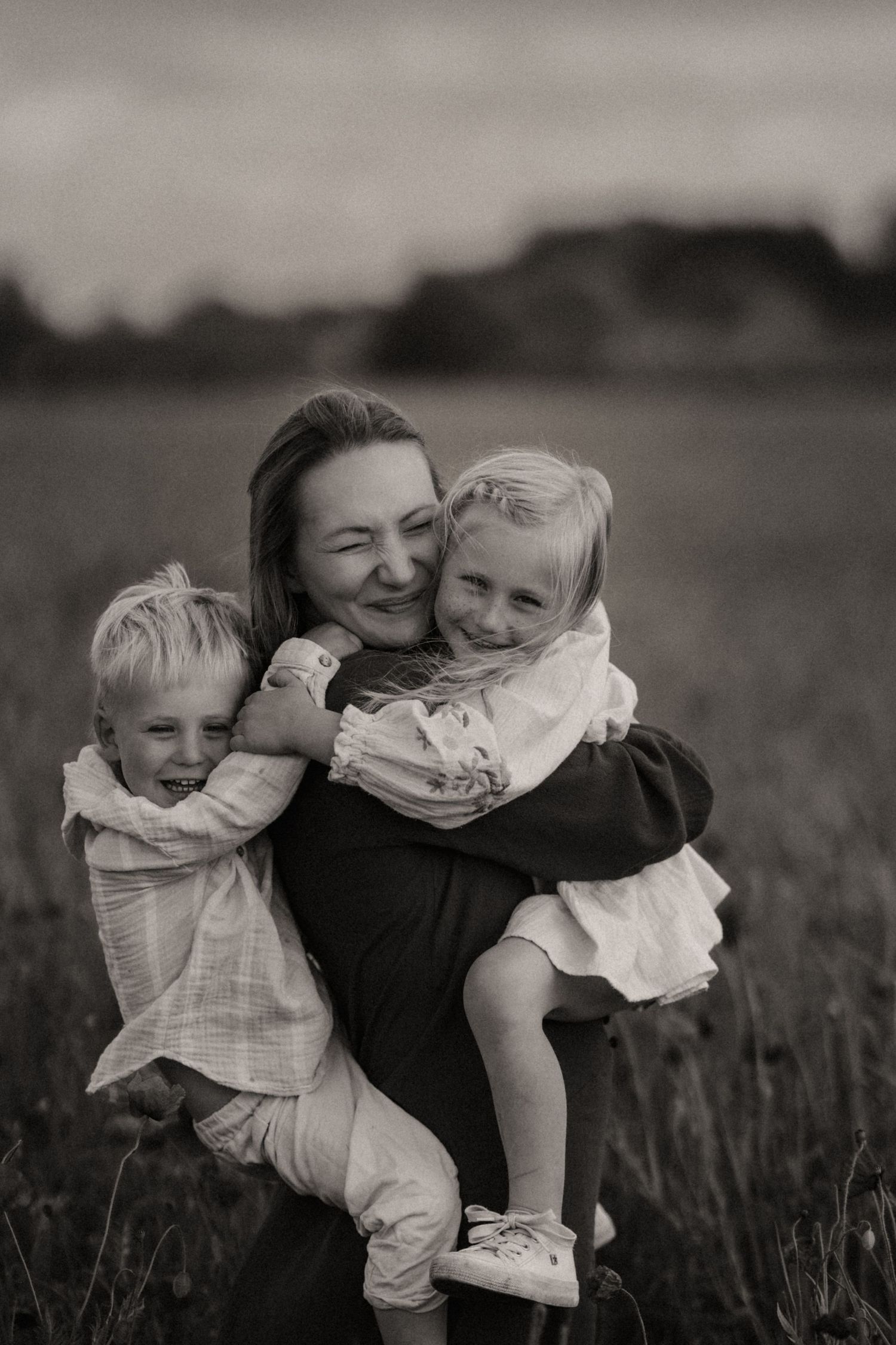 parent carrying two smiling children outdoors, hugging tightly during a candid black and white family photography session in Tamworth