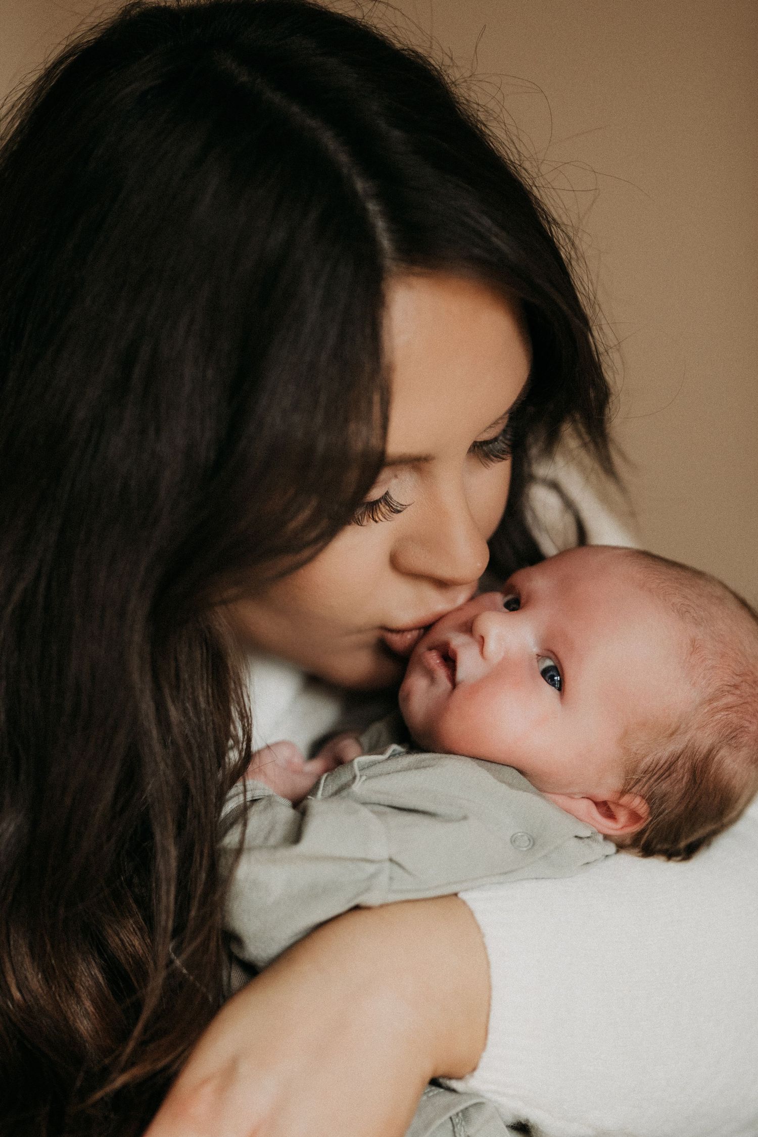 Mother gently kissing her newborn’s cheek while holding them close, a quiet moment captured during an intimate lifestyle newborn photoshoot.