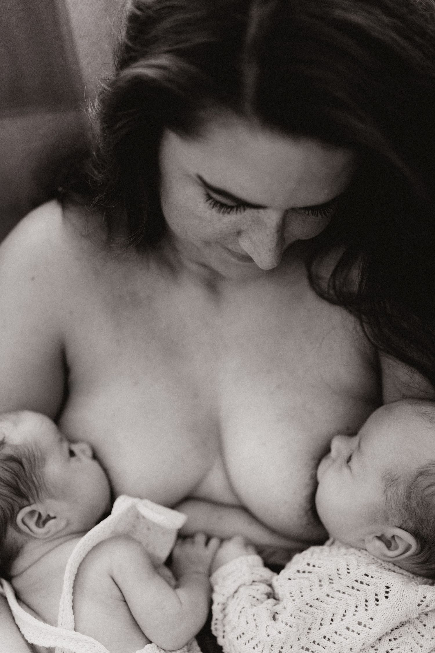 Mother looking down at her newborn twins as they rest against her chest, captured in a quiet, intimate moment of closeness