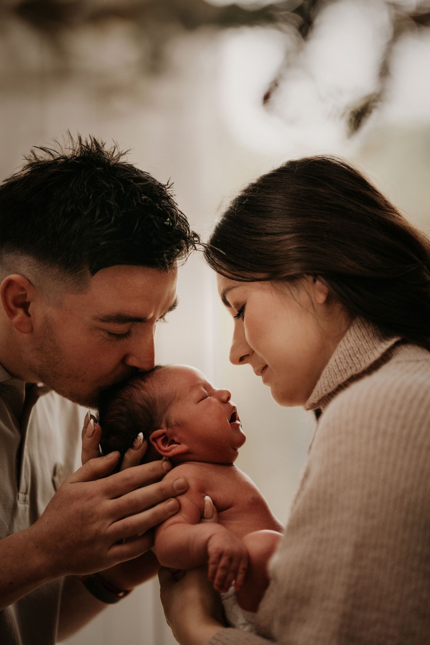 Parents hold their newborn close, sharing a kiss in soft golden light during a cozy family moment in Hinckley