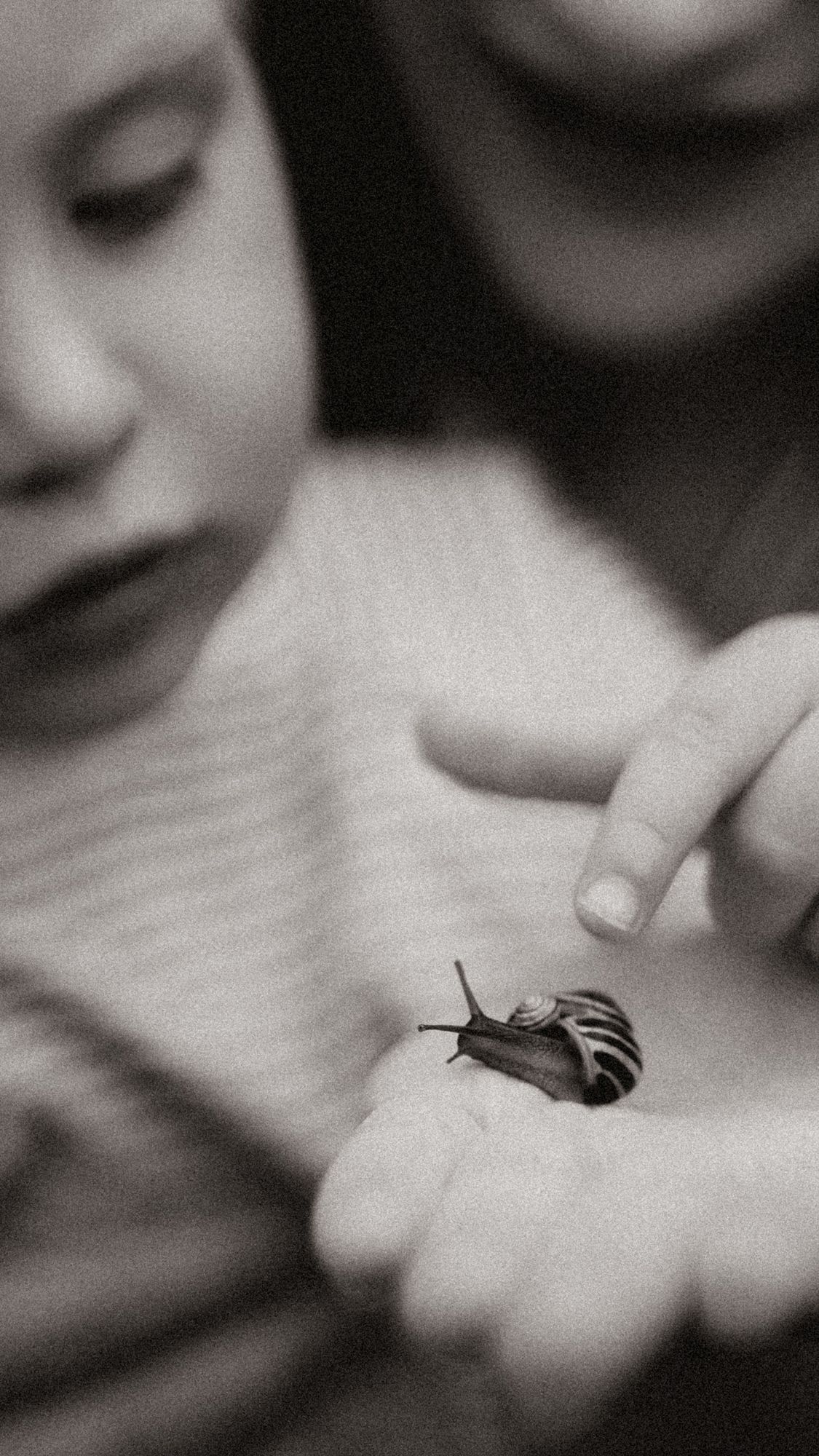 Child observing a tiny snail resting on a hand while a parent gently points to it, documented in a soft black and white lifestyle family ph