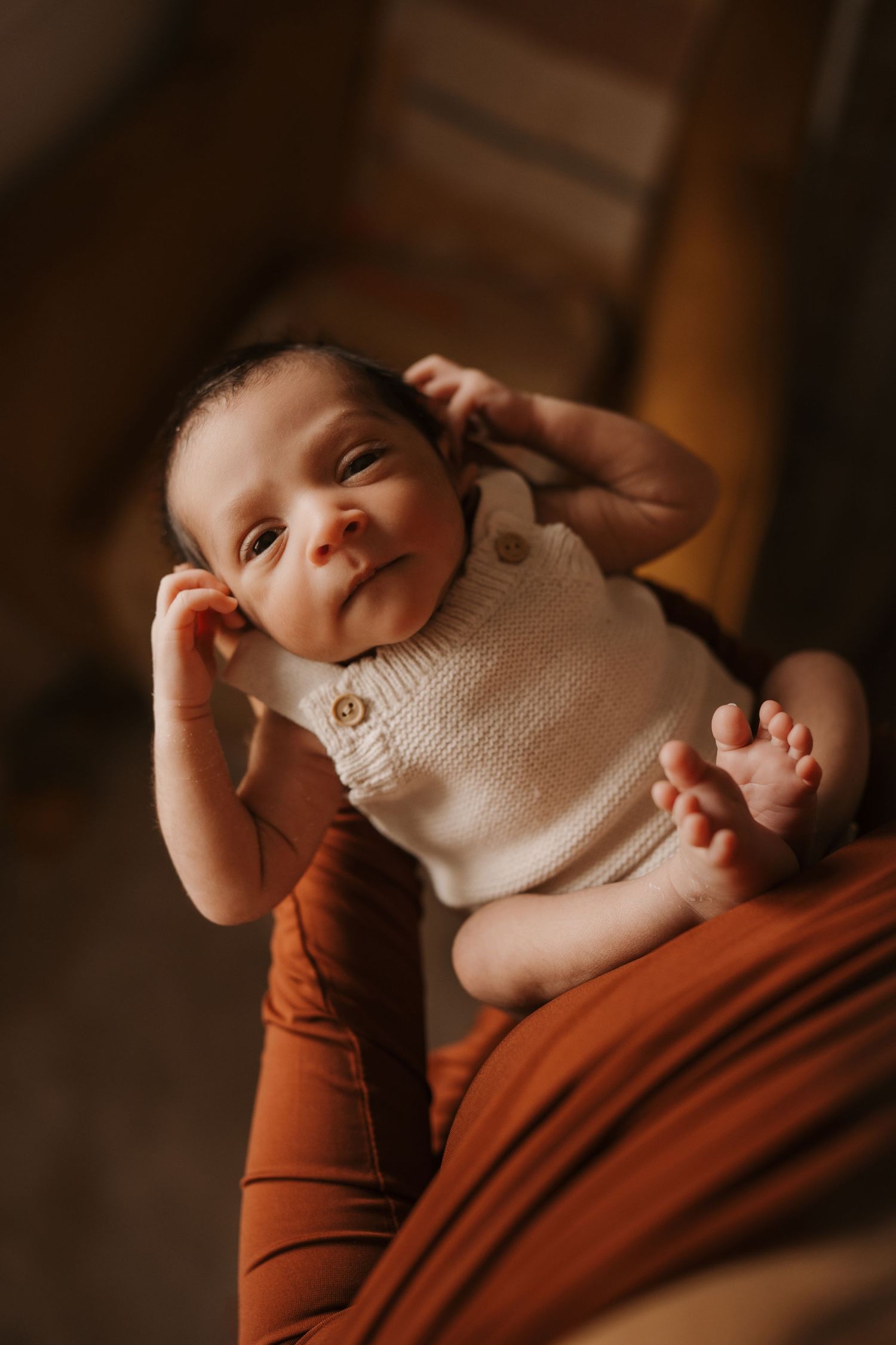 Newborn baby lying on a parent’s lap indoors, gently stretching arms and legs, wrapped in warm light and soft textures