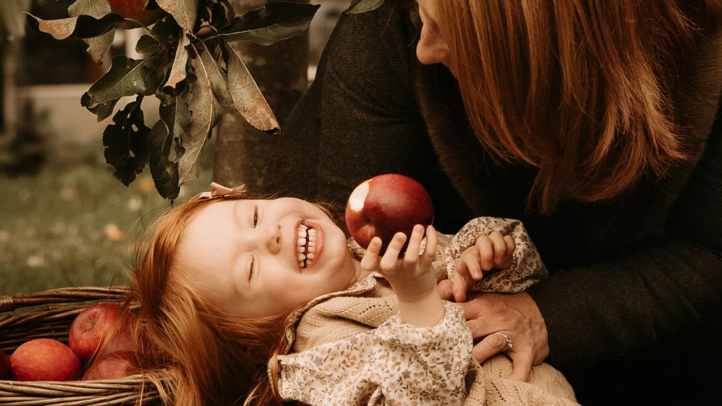 Laughing child lying back while holding an apple, surrounded by baskets of fruit and autumn tones, caught mid-giggle