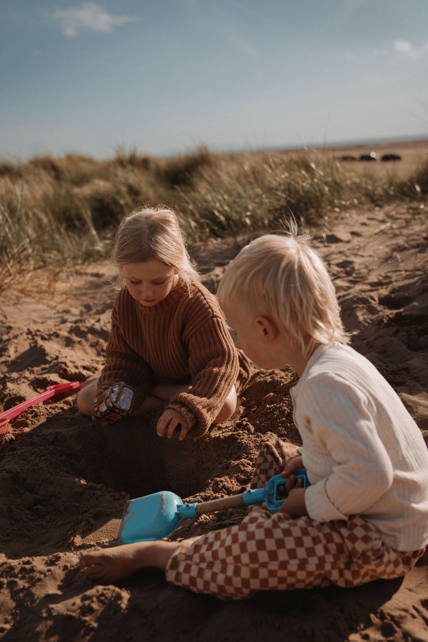 Two young children sitting in soft sand dunes, digging and playing with small shovels, heads bent together in quiet concentration under warm