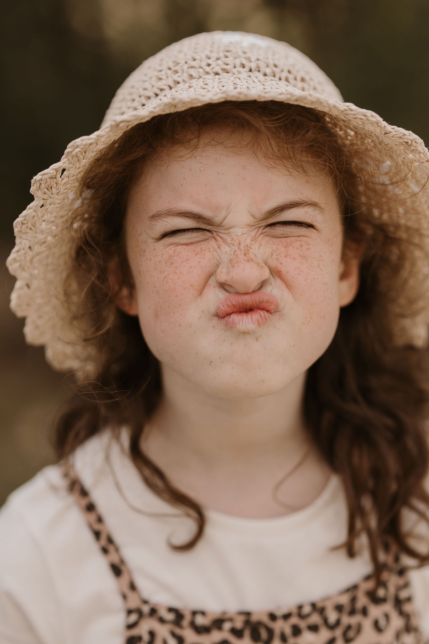 Young girl in a straw hat scrunches her face playfully, eyes closed and lips puckered, standing outdoors in soft natural light.