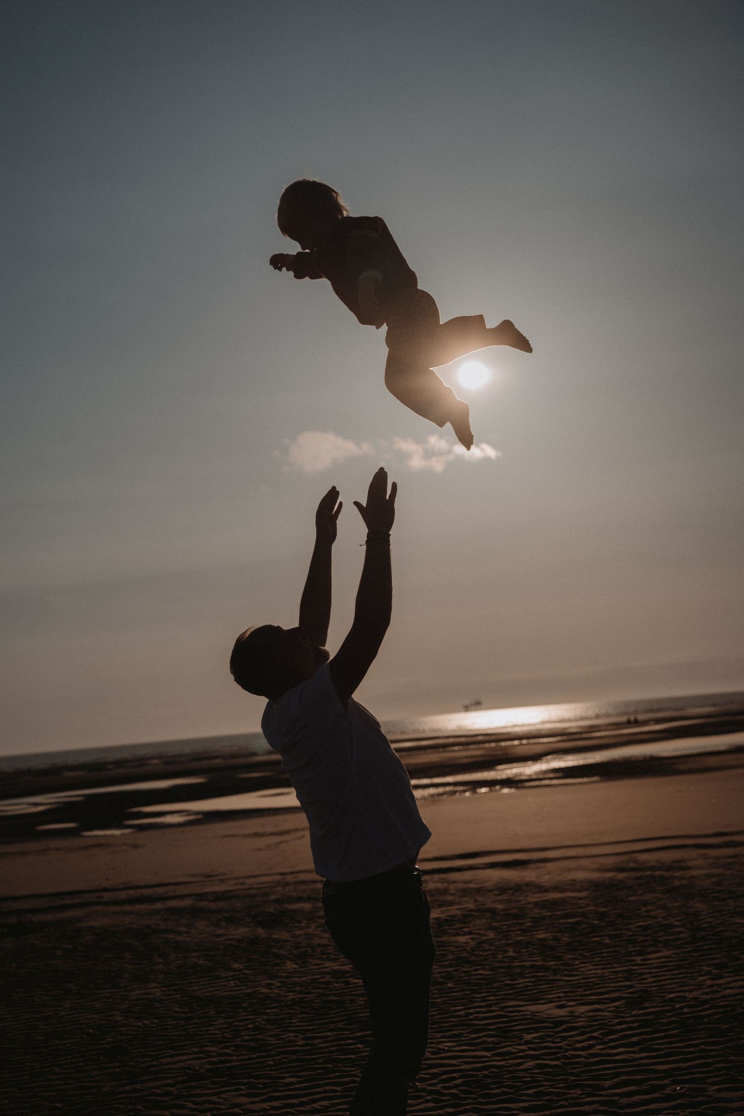 Silhouette of a parent lifting a child high into the air on a beach at sunset, the child floating joyfully against the glowing sky and horiz