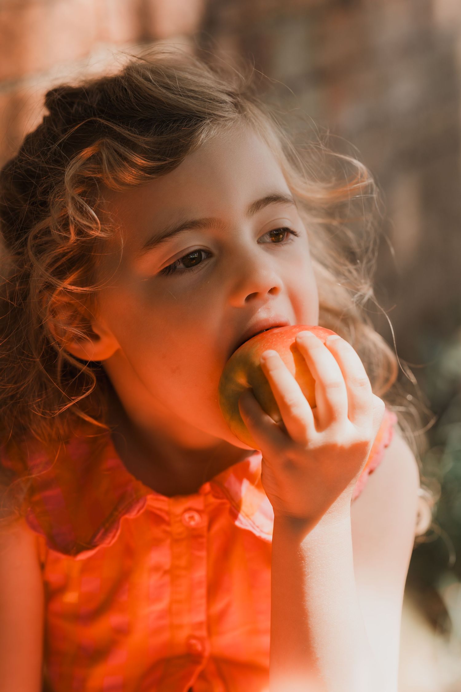 Little girl in orange dress eats an apple during a natural, outdoor childhood photoshoot