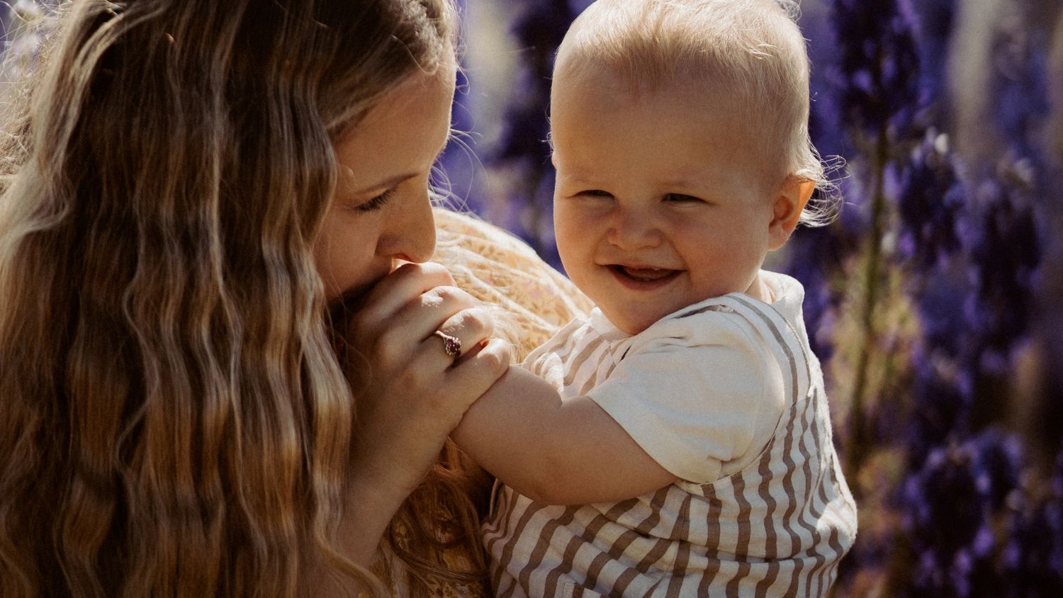 Mother holding a smiling baby outdoors among purple flowers, sharing a quiet moment as the baby looks toward the camera