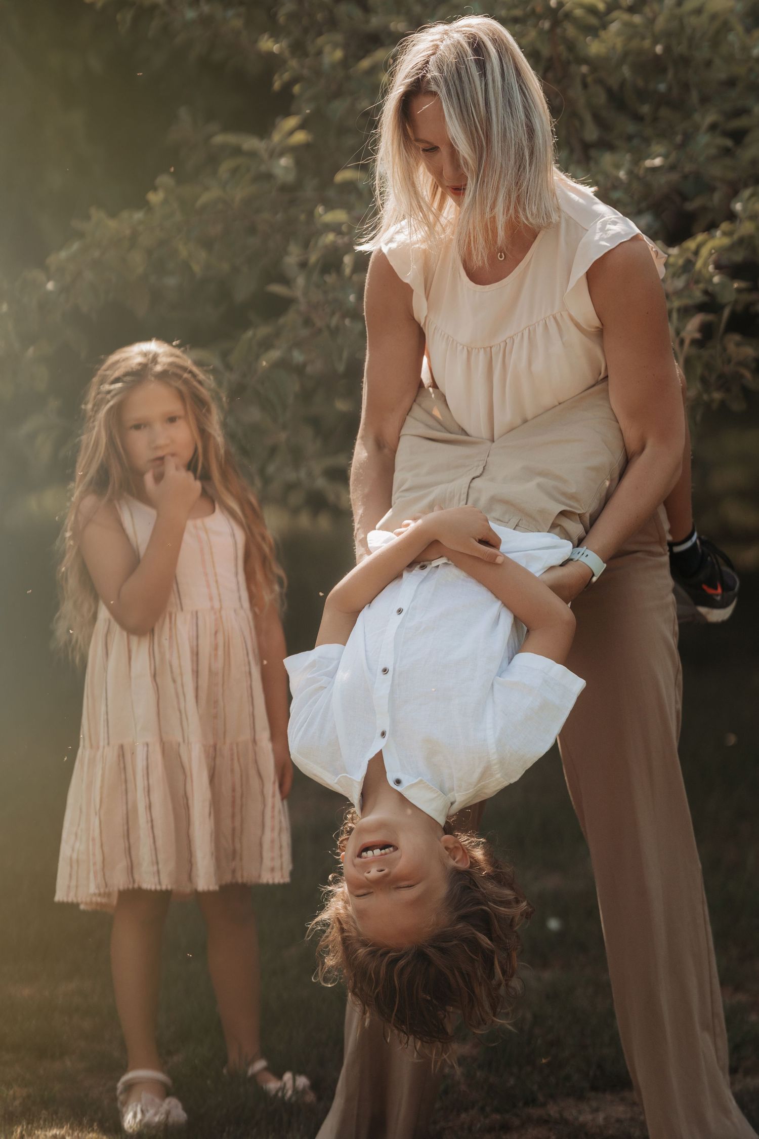 Mother standing outdoors with two young children, one child held upside down playfully while the other watches with curiosity
