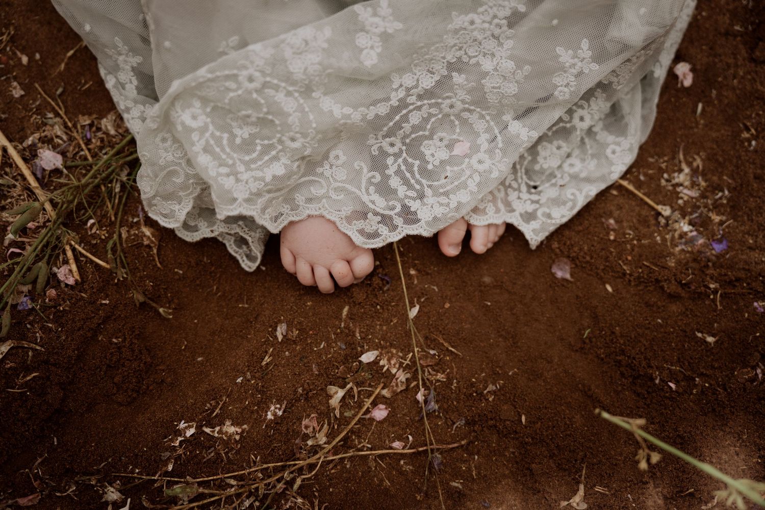 Baby's Bare feet in a lace dress standing on earthy ground, captured during a natural Confetti Field outdoor storytelling photoshoot.