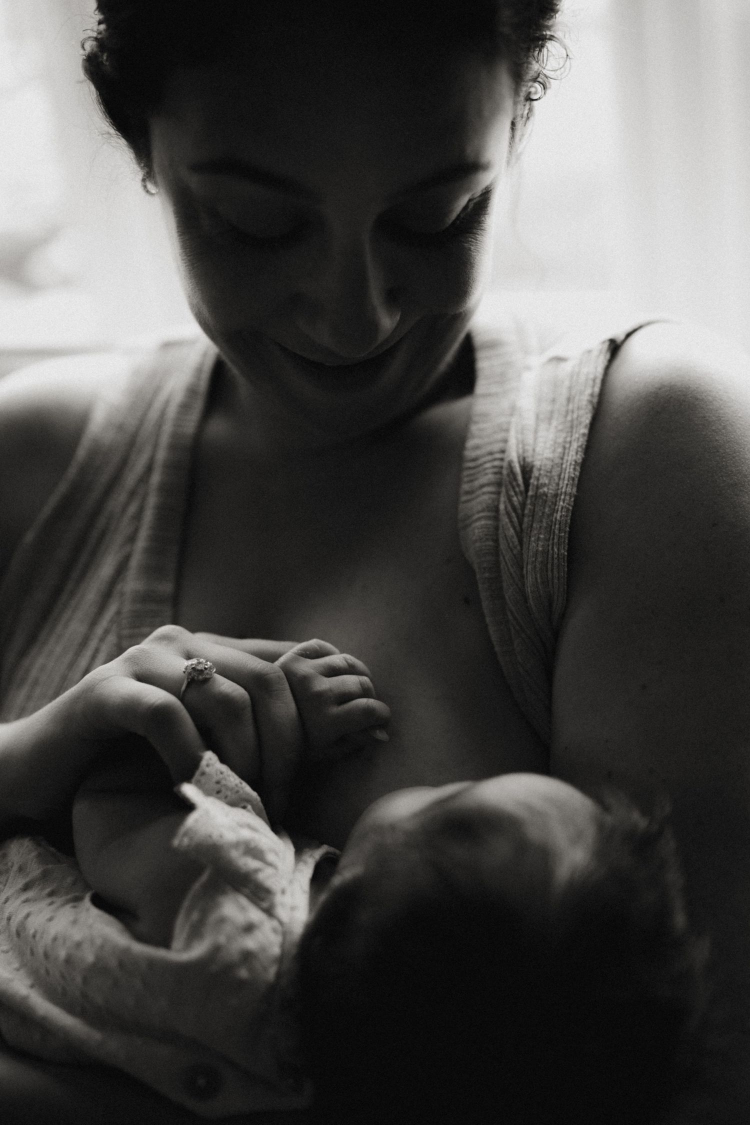 Mother nurses her newborn by the window, smiling softly in a peaceful black-and-white portrait.