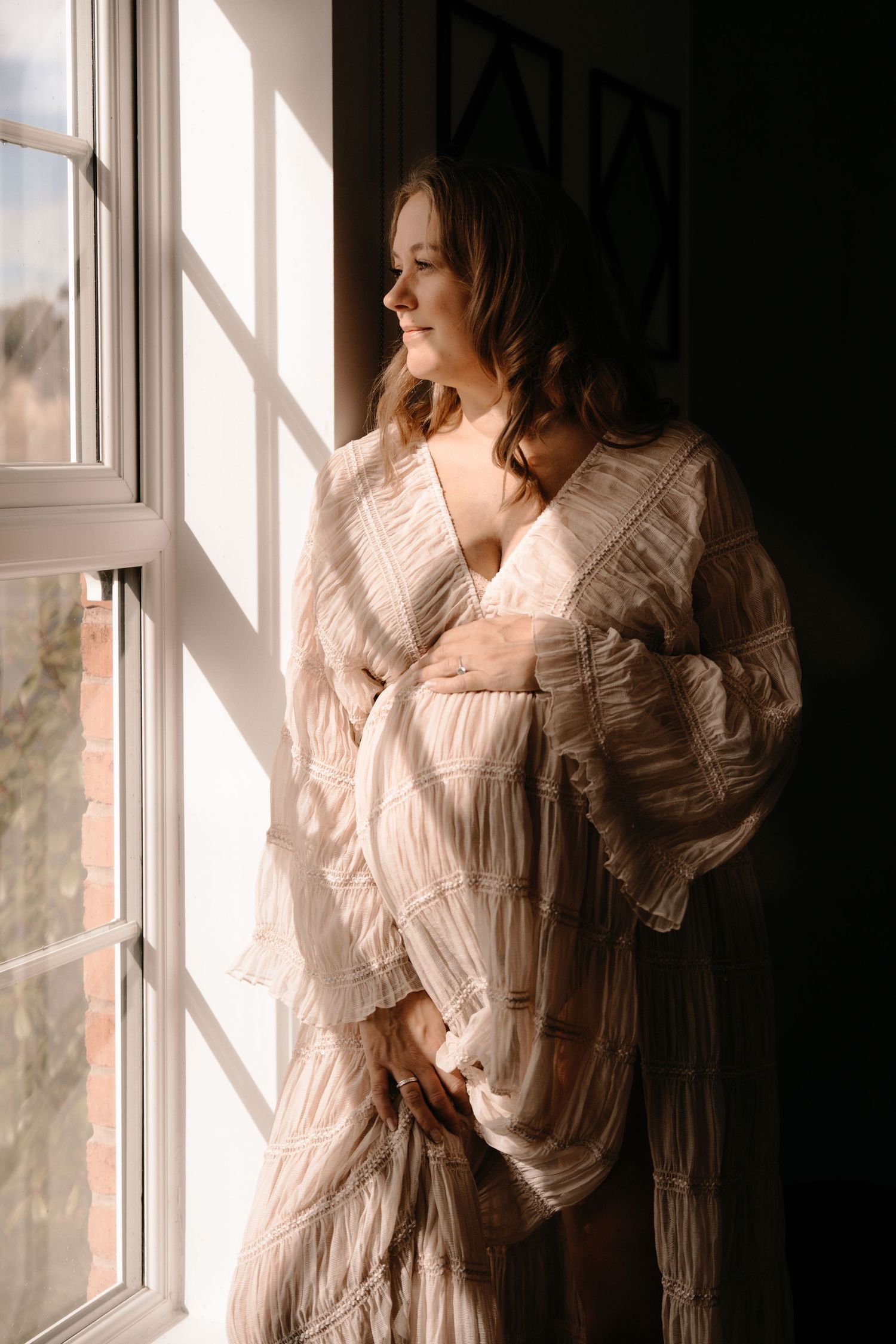 Pregnant parent standing by a window in soft light, wrapped in a cozy cardigan during a relaxed maternity session.